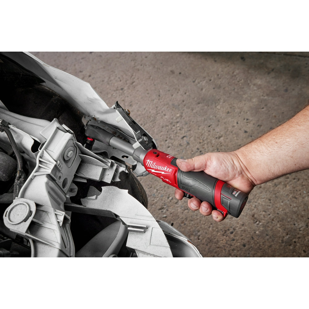 Hand holding red and black Milwaukee M12 FUEL ratchet with protective boot, positioned near white work light on concrete surf