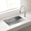 Stainless steel single bowl sink with faucet mounted on white countertop near window.