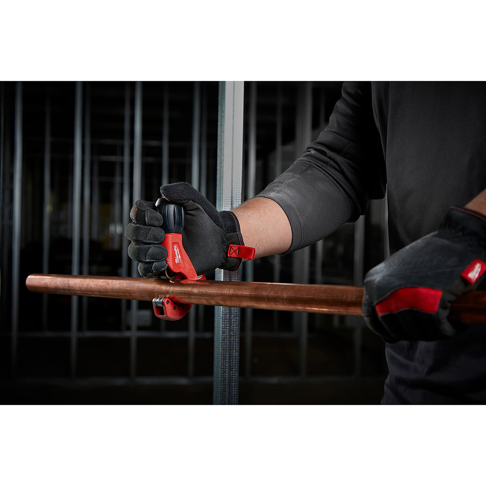 Red and black tubing cutter positioned on copper pipe, held by gloved hands.