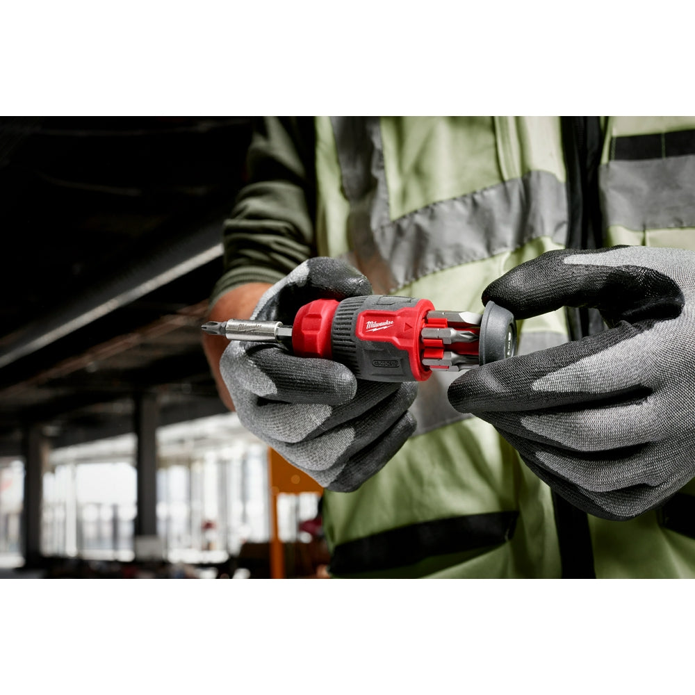 Red and black compact multi-bit screwdriver with storage slots held in gloved hands at a job site.