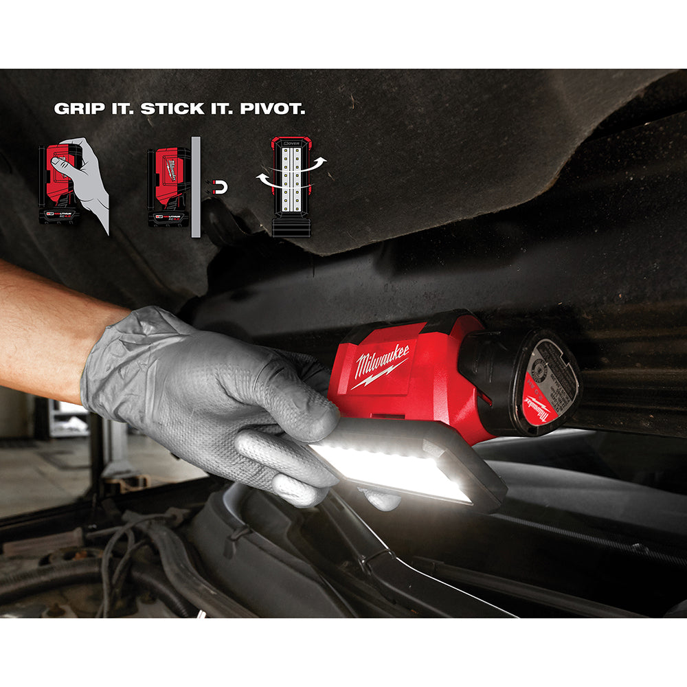 Red and black Milwaukee M12 flood light with bright white LED head held in gloved hand under vehicle, showing pivot attachmen