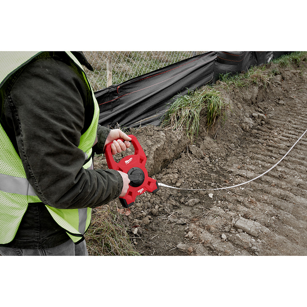 Worker in safety vest holding red open reel tape measure with extended steel tape across dirt ground.