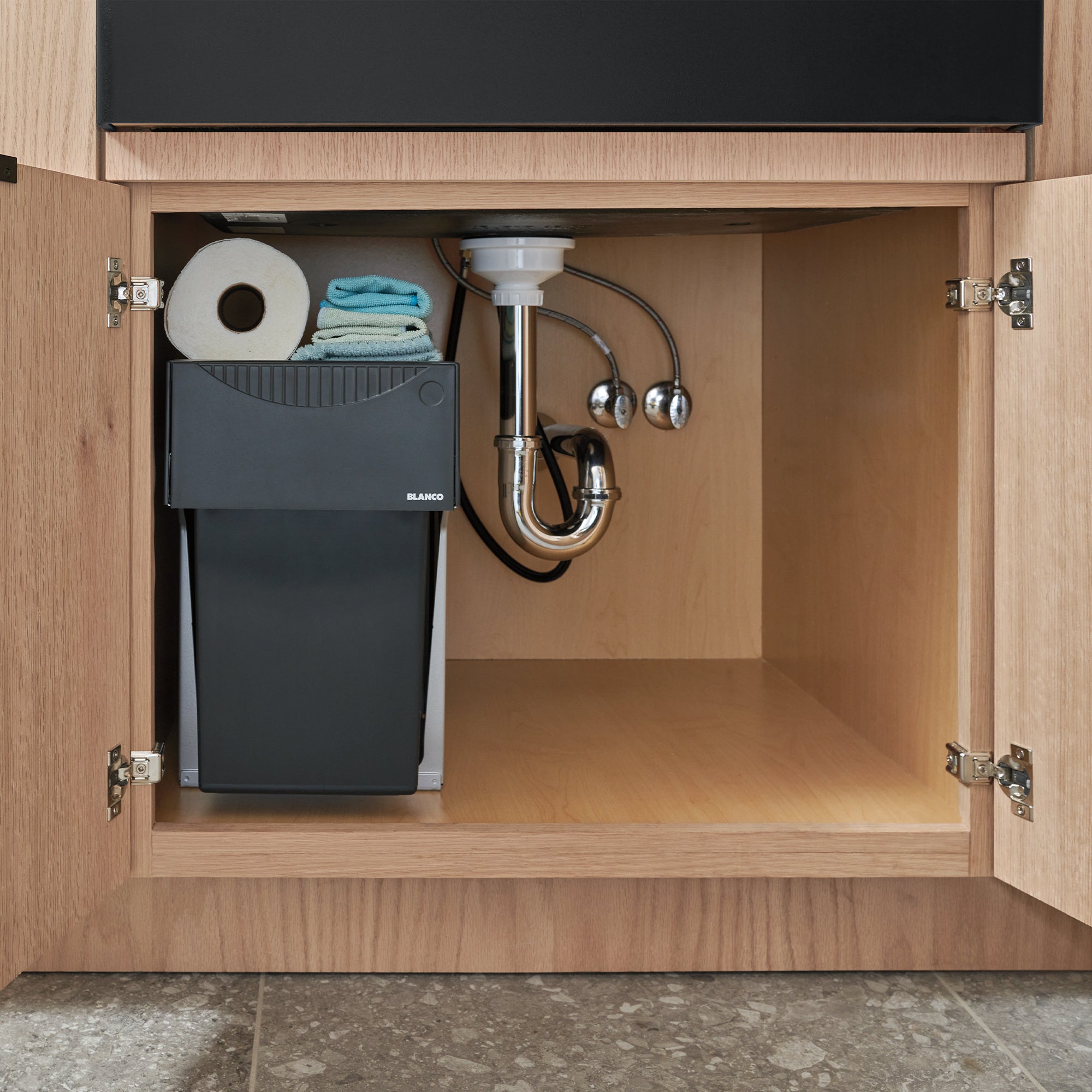 Under-sink cabinet with black trash bin, chrome P-trap plumbing, and compartment storing paper towels and blue cloths.