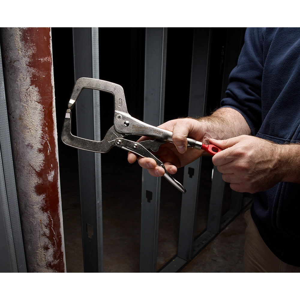Metal C-clamp with locking mechanism and red handle held in hand, clamping a rusty metal rod.