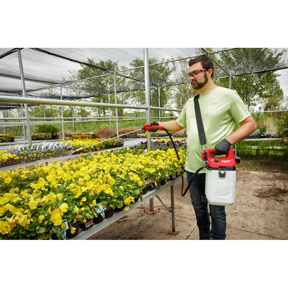 Black shoulder strap attached to red and white handheld sprayer worn across person's torso in greenhouse setting.