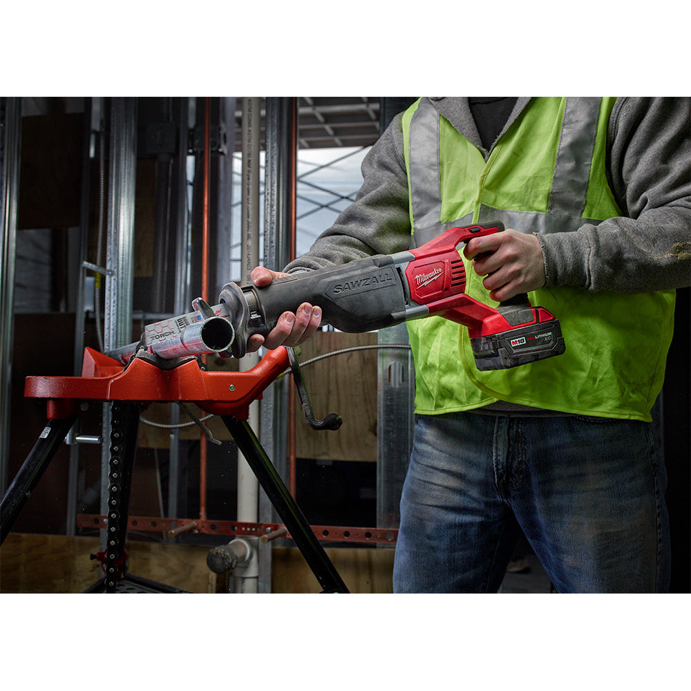 Red and black Milwaukee M18 reciprocating saw held by worker in safety vest, cutting metal pipe on work vise.