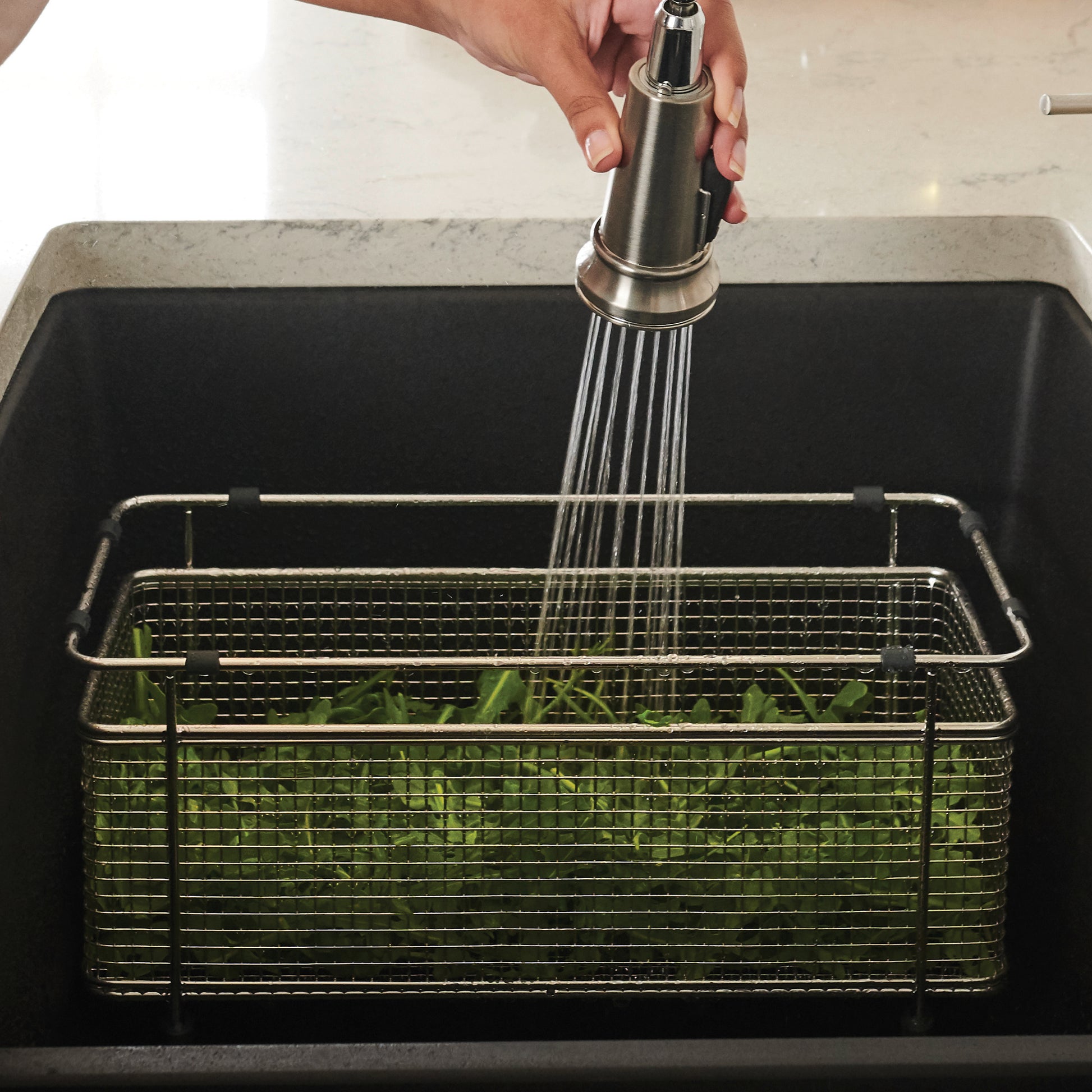 Stainless steel mesh colander basket with handles sitting in sink while faucet sprays water over fresh greens inside.