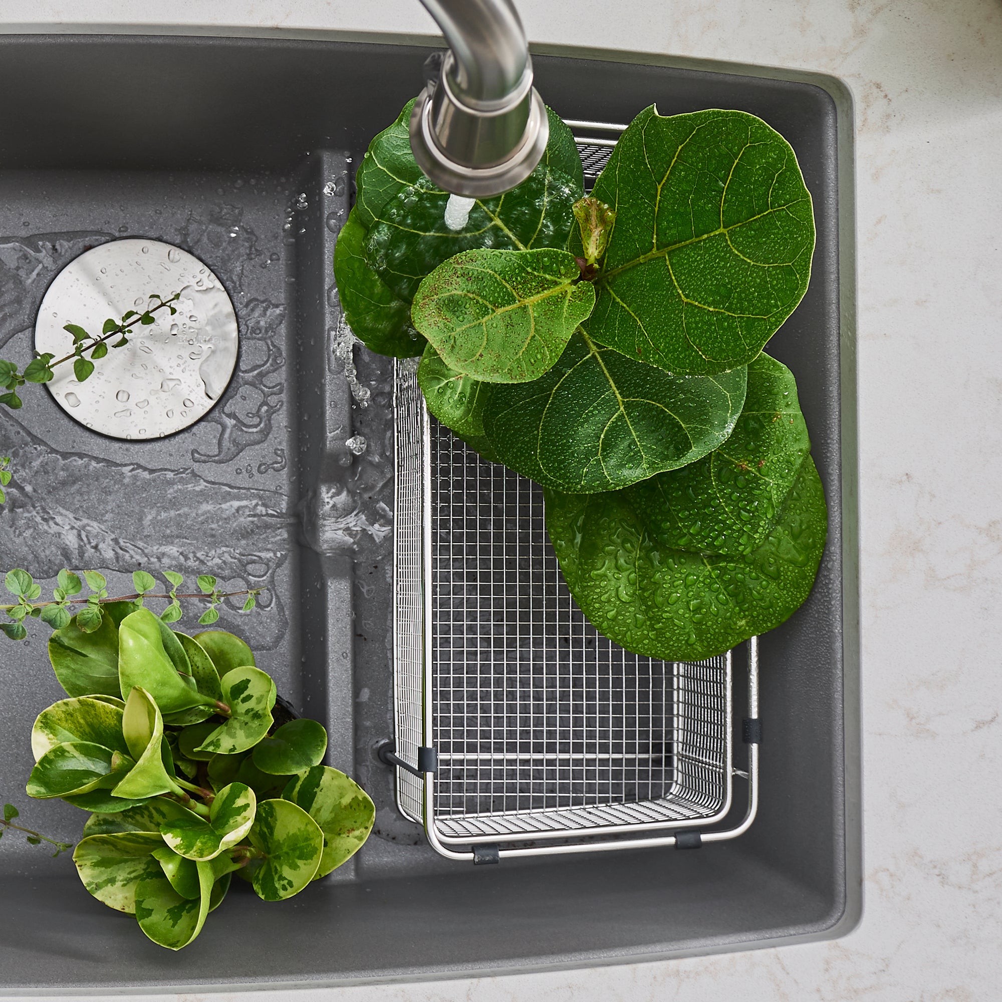 Stainless steel mesh colander with fine grid pattern positioned in dark sink basin with water and fresh greens.