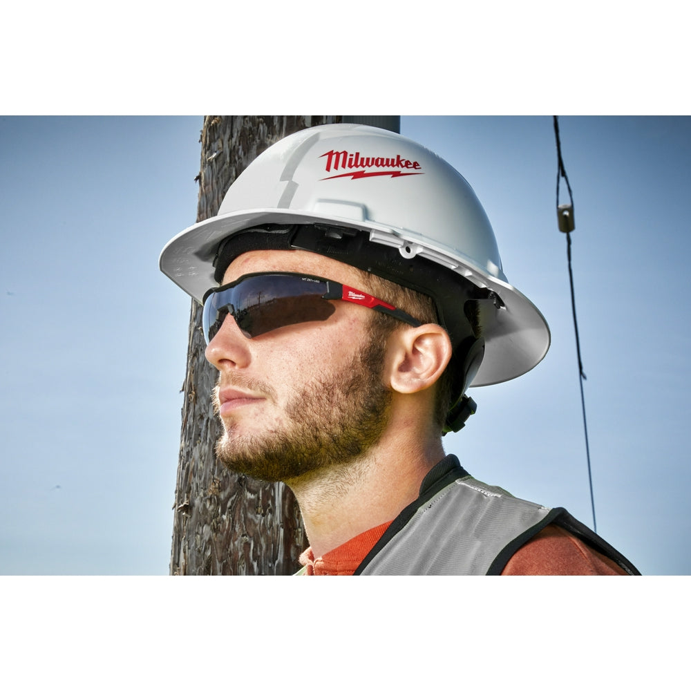 Milwaukee safety glasses with tinted dark lenses and red accents worn by worker in hard hat.