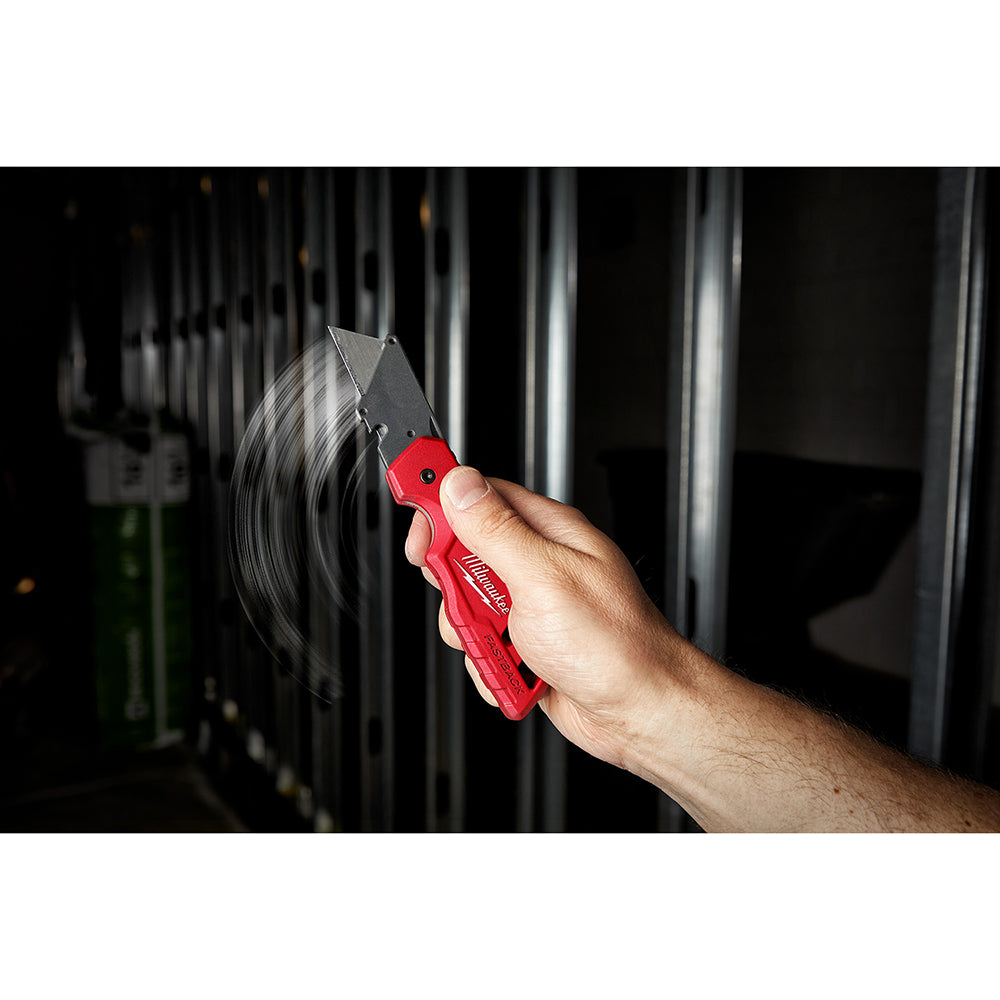 Red and black folding utility knife with blade extended, held in hand near metal fencing.