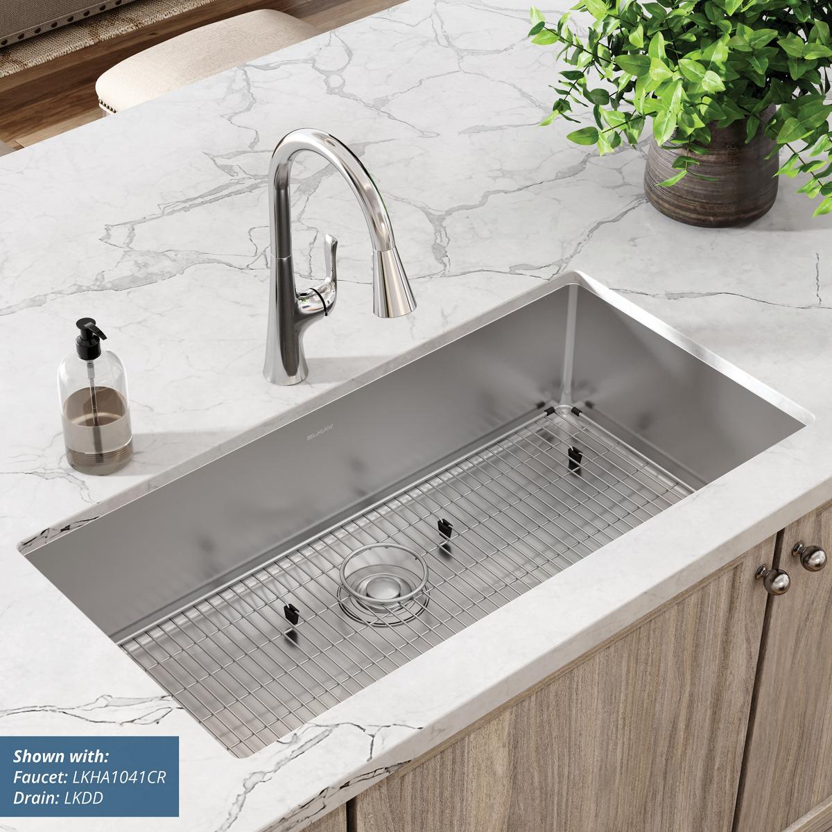Stainless steel single bowl undermount sink installed in white marble countertop with metal grid insert and faucet.