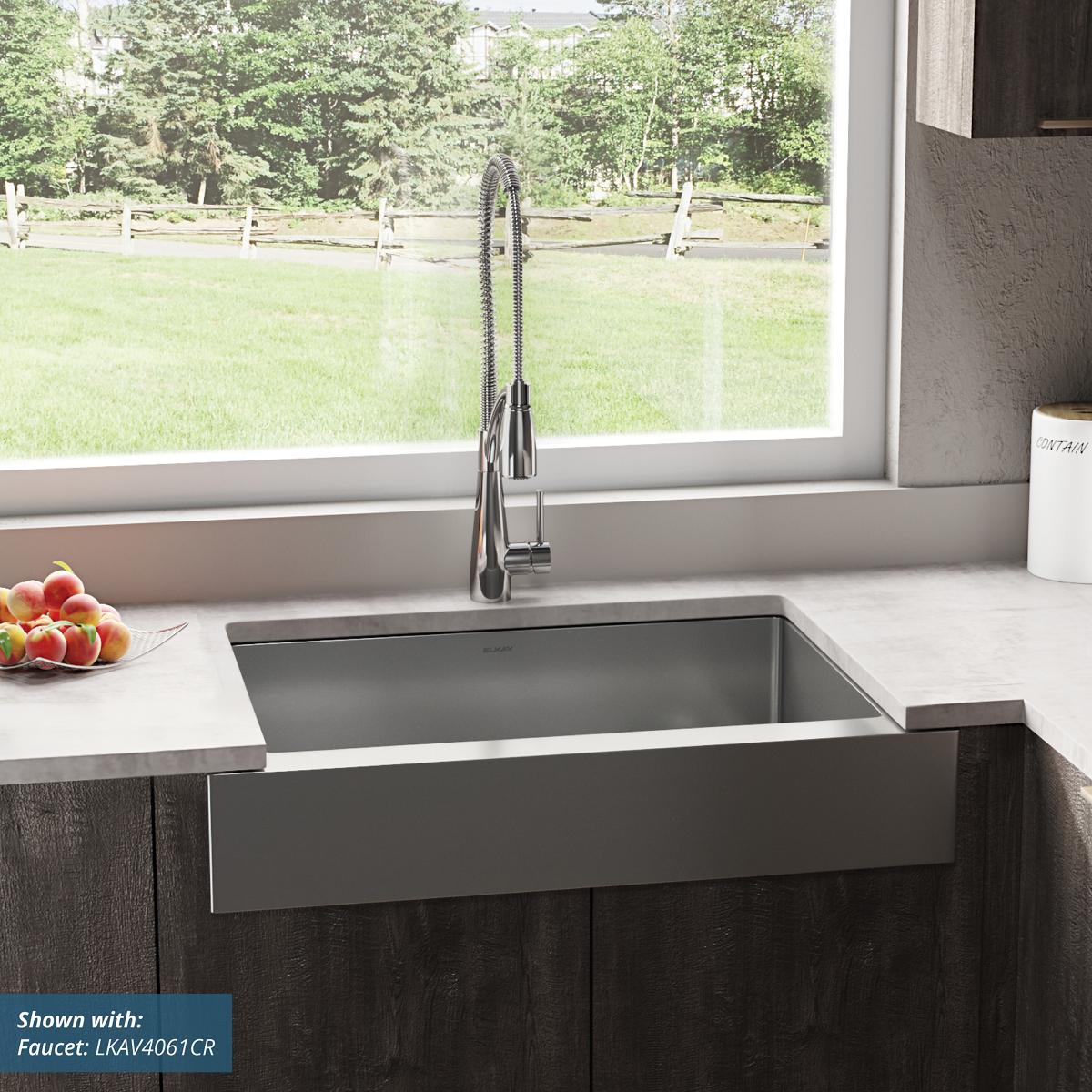 Stainless steel farmhouse sink with single bowl and apron front installed in dark cabinetry with pull-down faucet.