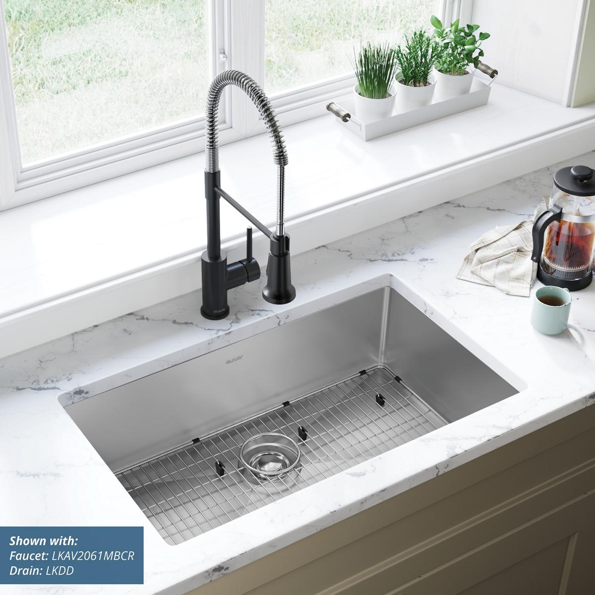 Stainless steel undermount sink with single bowl, grid, and drain strainer installed in white countertop with black faucet.