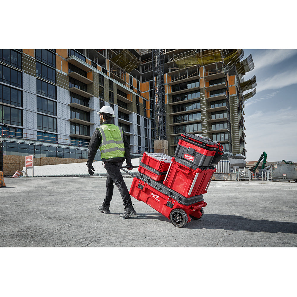 Red and black modular tool storage boxes stacked on wheeled base being transported by worker on construction site.