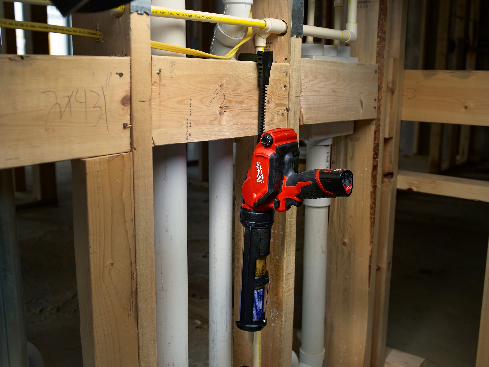 Red and black Milwaukee caulking gun with aluminum barrel mounted on wooden framing structure.