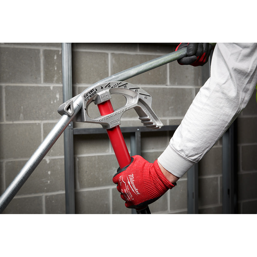 Milwaukee aluminum conduit bender with red handle held in gloved hands against brick wall background.