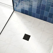 Matte black square floor drain installed in white marble tile with blue tile wall visible in background.