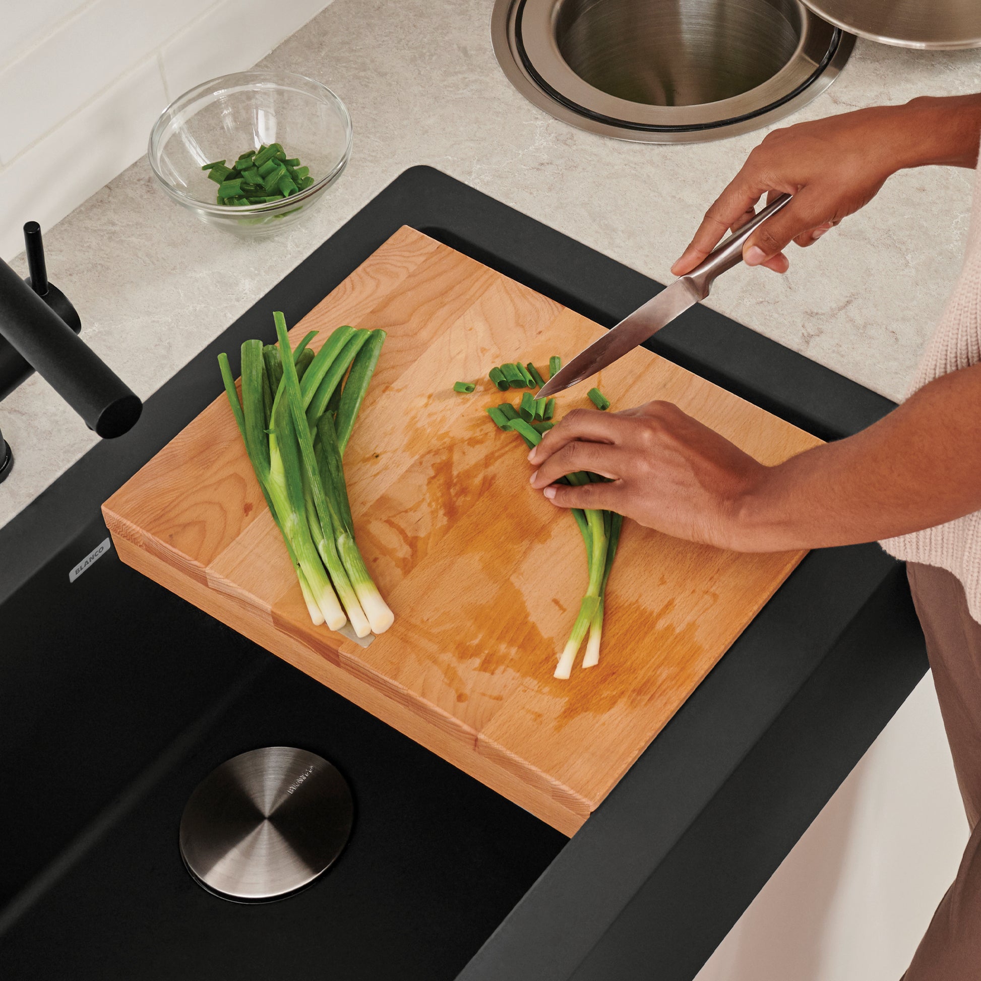 Beechwood cutting board with black trim resting across apron front sink while person chops green onions.