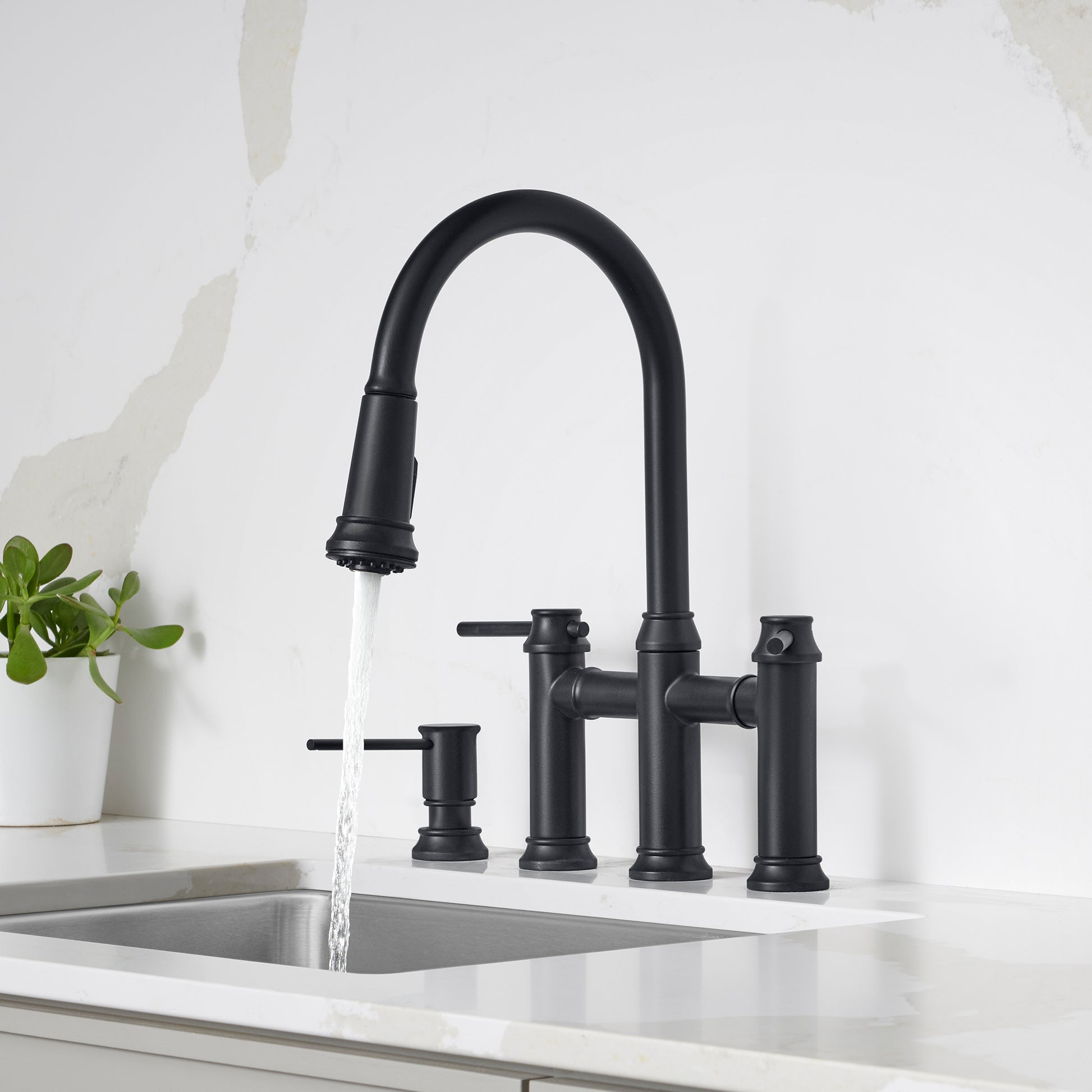 Matte black pull-down bridge faucet with curved spout, lever handles, and soap dispenser on white countertop.