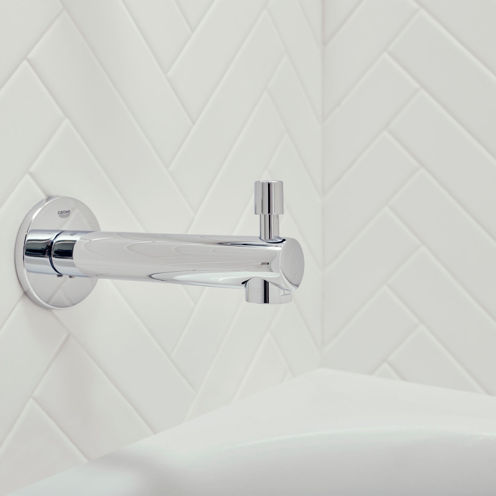 Chrome tub spout with diverter lever mounted on white herringbone tile wall above bathtub.