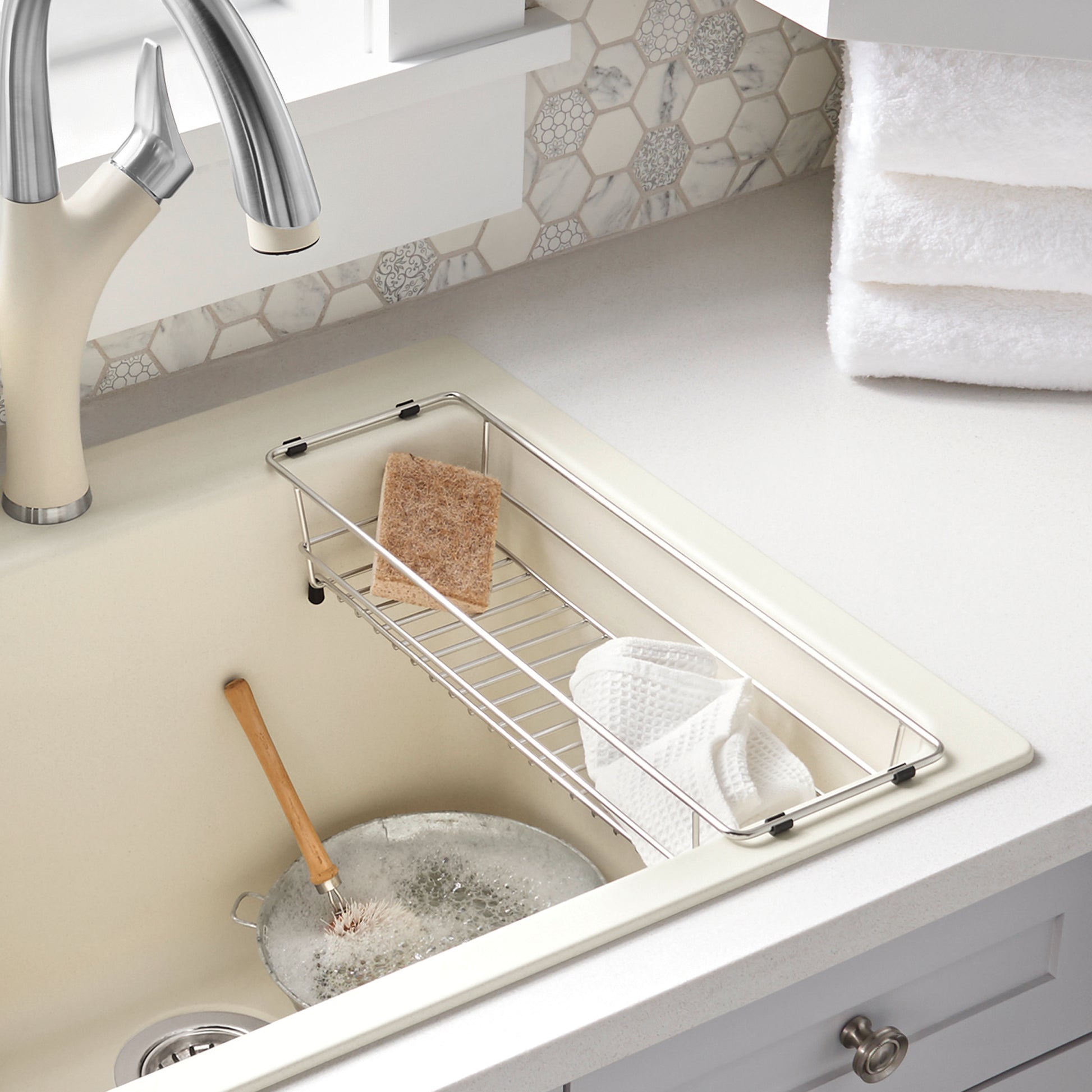 Stainless steel rectangular basket with grid pattern holding a brown sponge and white cloth inside a laundry sink.