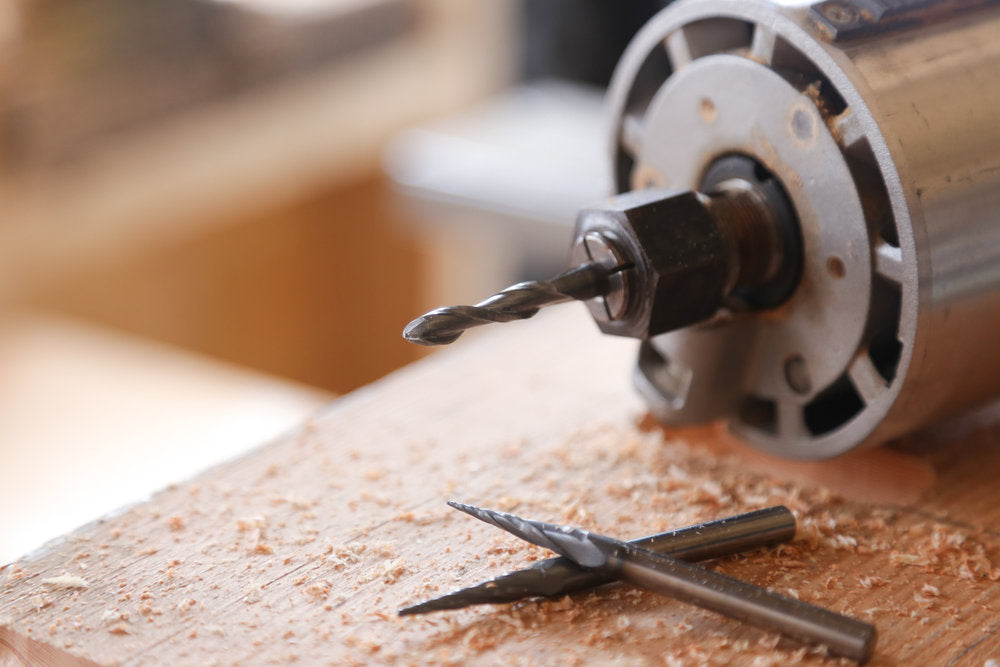 Three metal router bits positioned next to a mounted CNC router with wood shavings on workpiece.