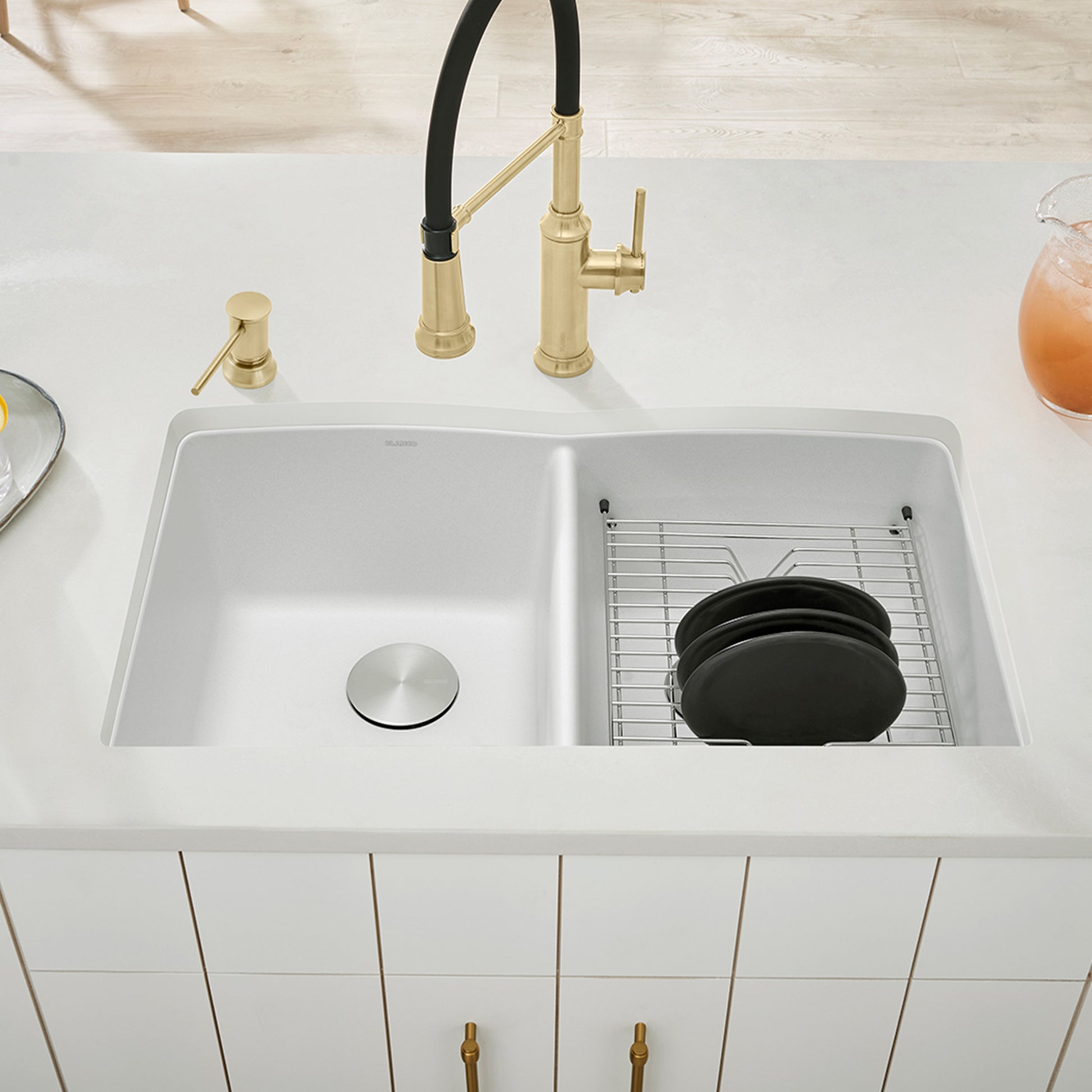 White double bowl undermount kitchen sink with metal drain plug and grid insert, paired with brass faucet and black handle.