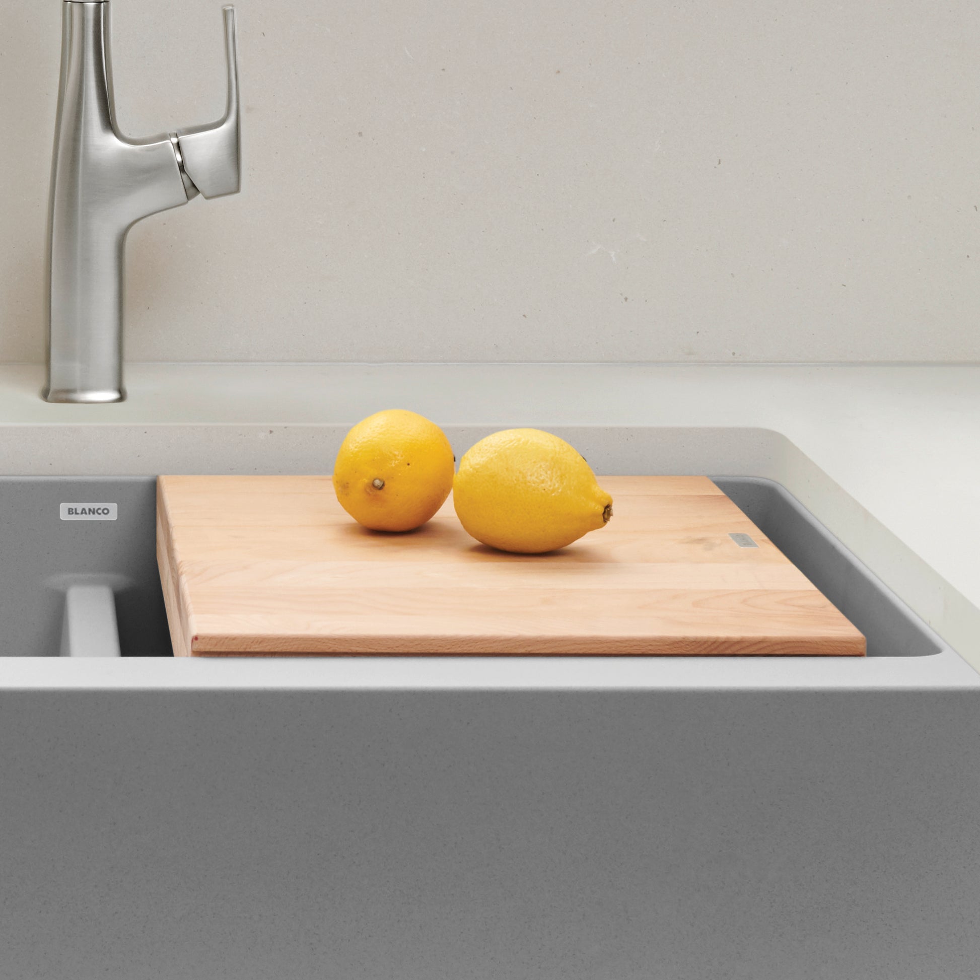 Beechwood cutting board spanning across gray apron front sink with two lemons on surface and faucet visible.