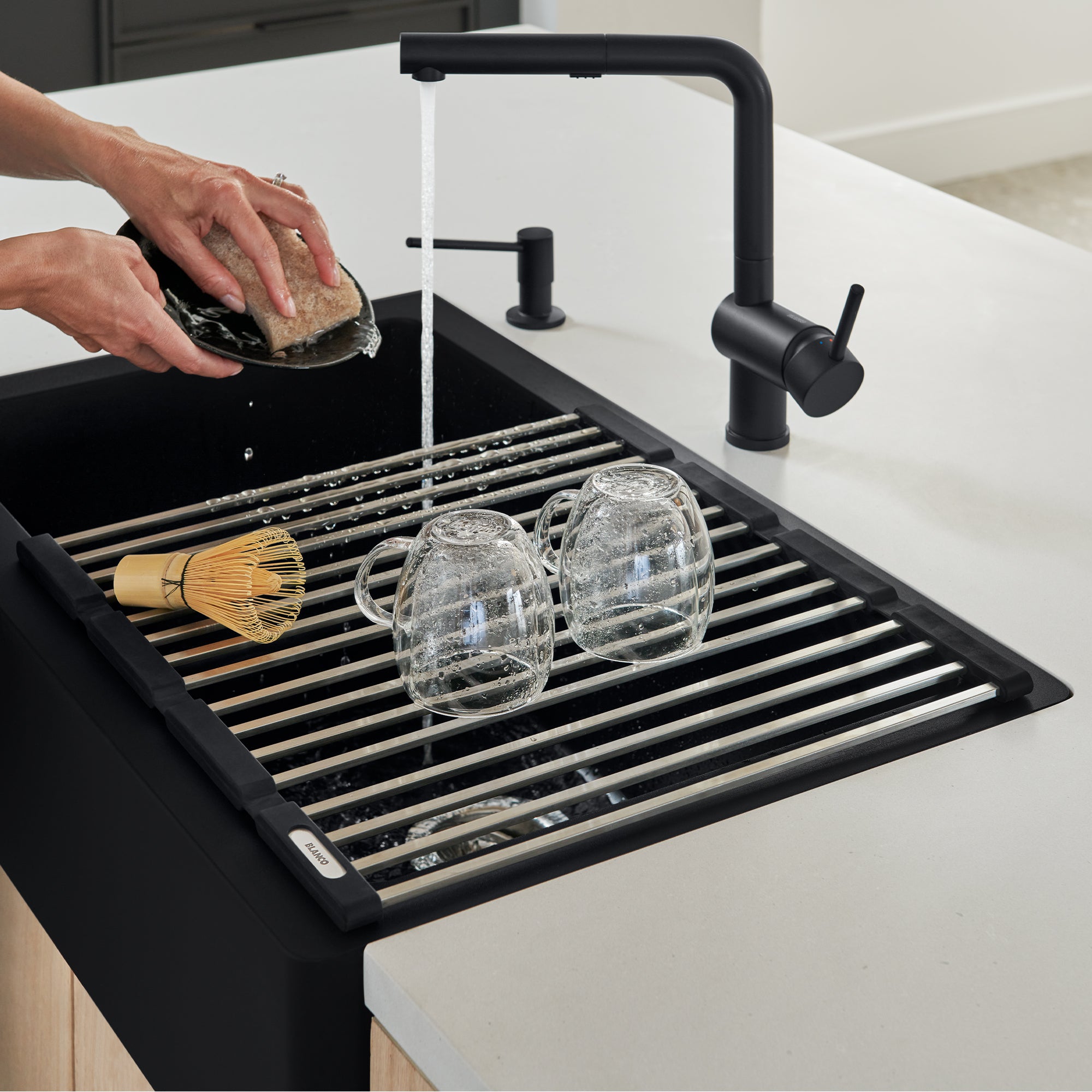 Black foldable drying mat with parallel rods inside kitchen sink, holding glasses and brush while hand rinses sponge under fa