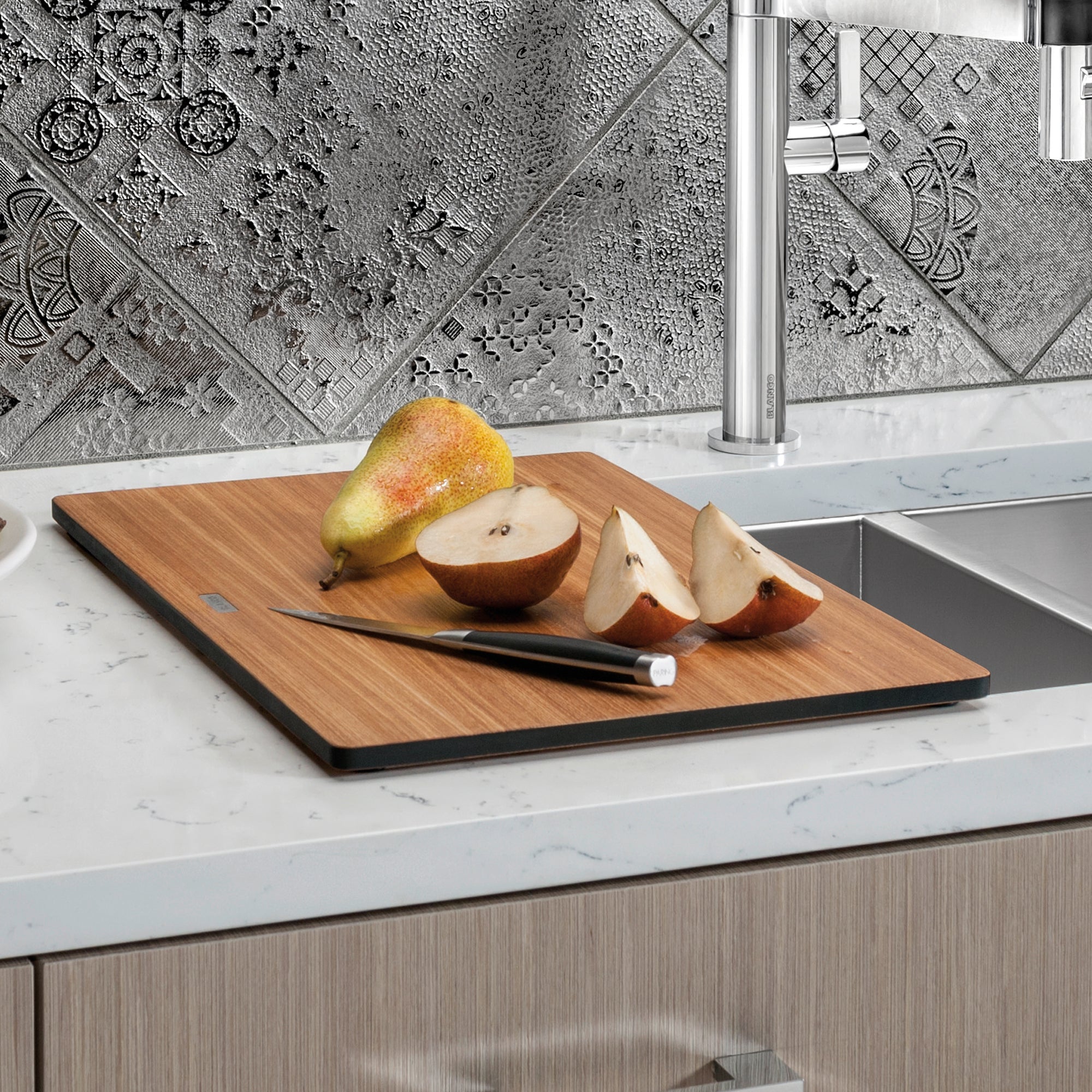 Rectangular wooden cutting board with black border containing sliced pear and apple with knife on white kitchen counter.