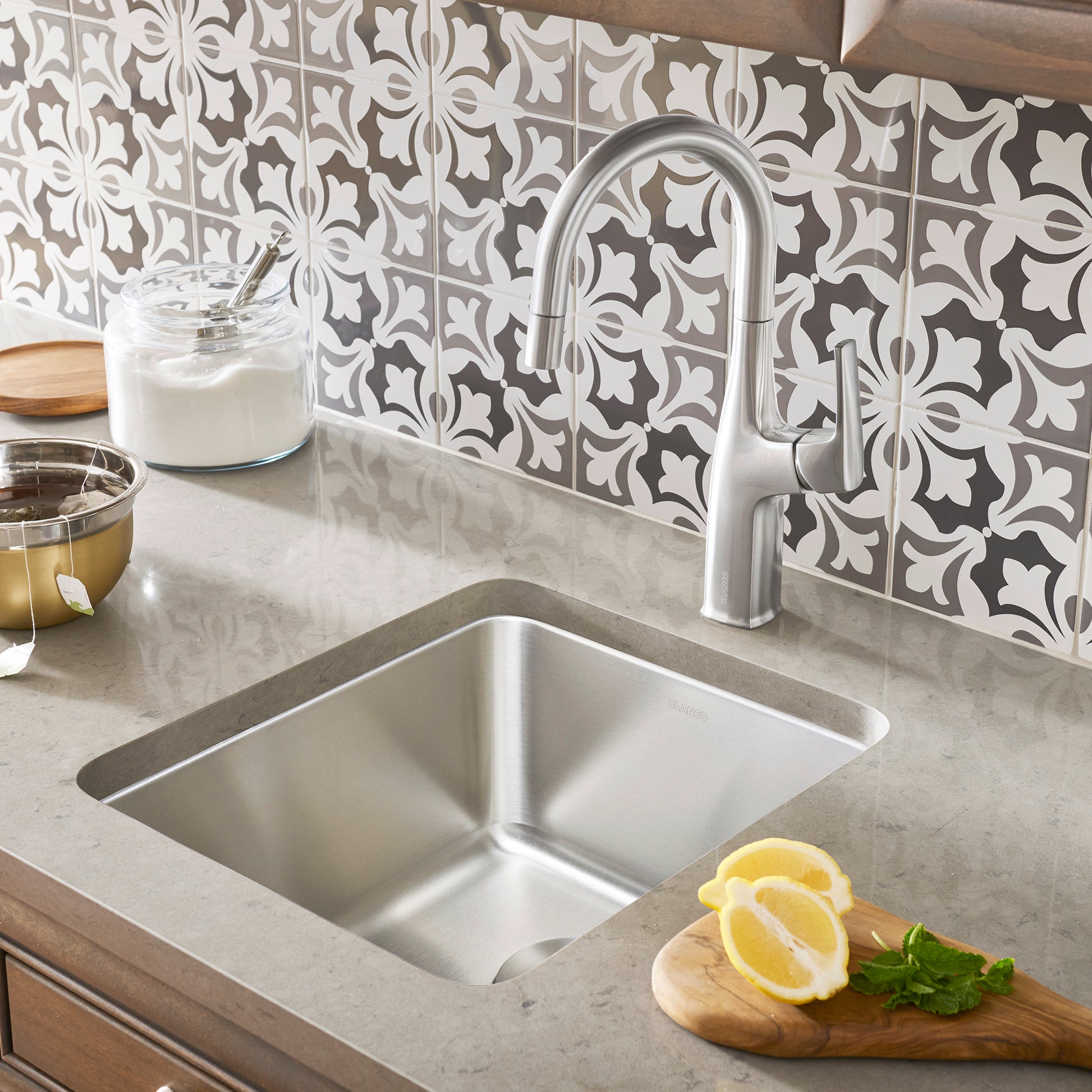 Stainless steel undermount bar sink installed in countertop with chrome arc faucet and patterned tile backsplash.