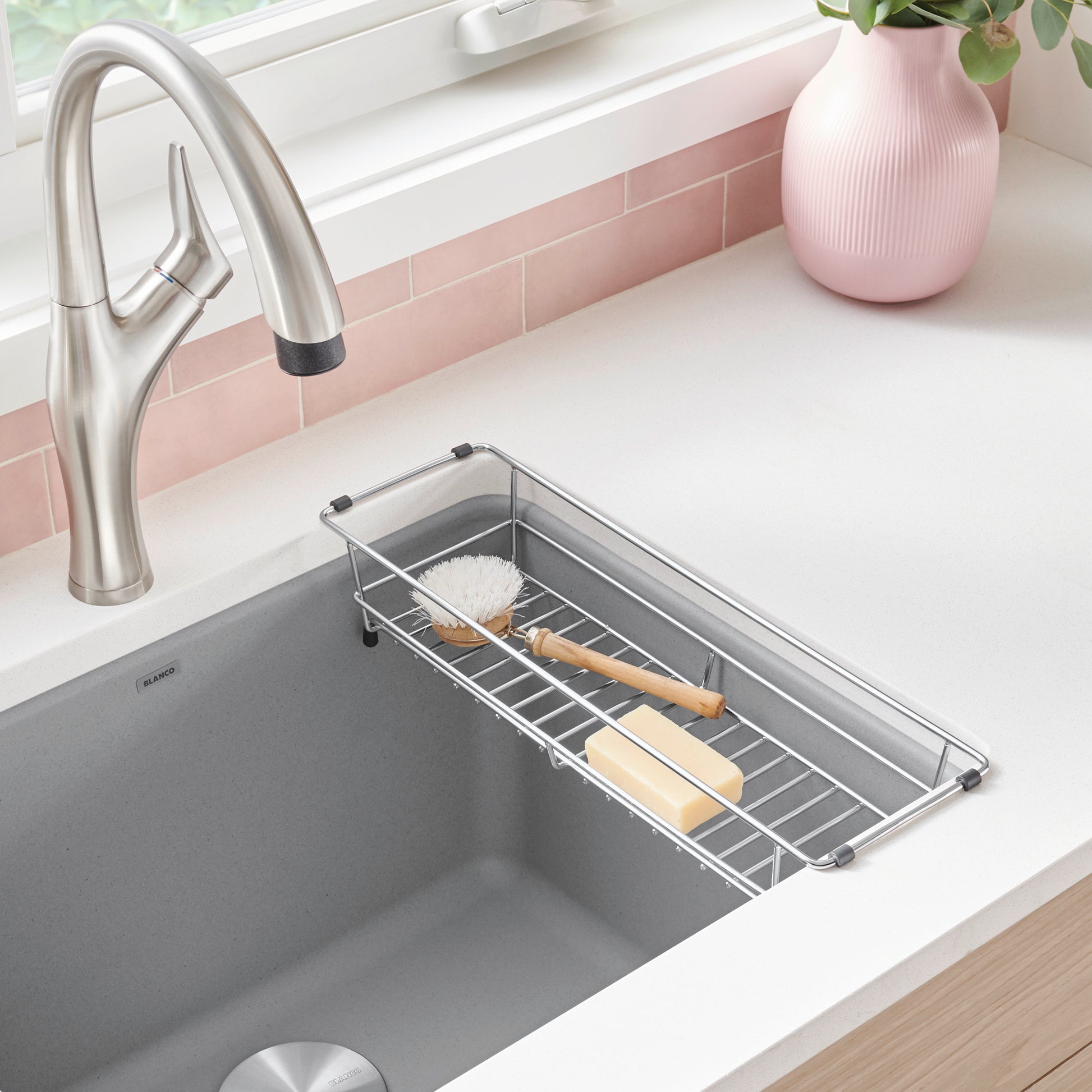 Stainless steel sink basket with slotted design holding brushes and utensils, positioned across laundry sink.