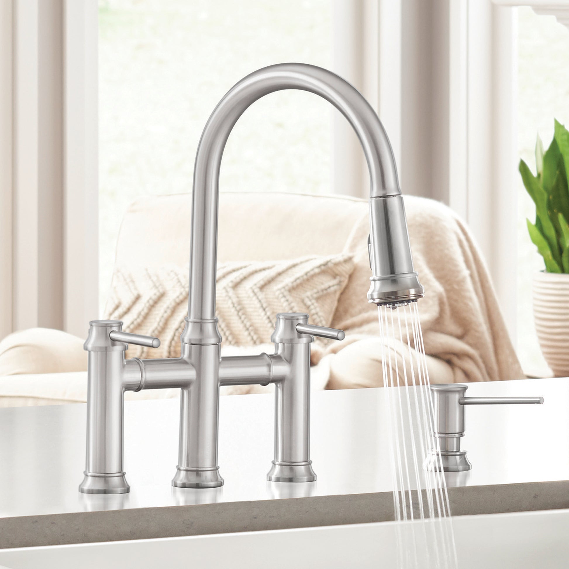 Stainless steel bridge faucet with curved pull-down spout, dual handles, and side sprayer on kitchen sink.
