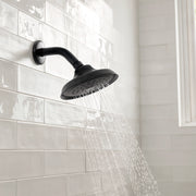 Matte black fixed shower head with curved arm mounted on white subway tile wall, water spraying downward.