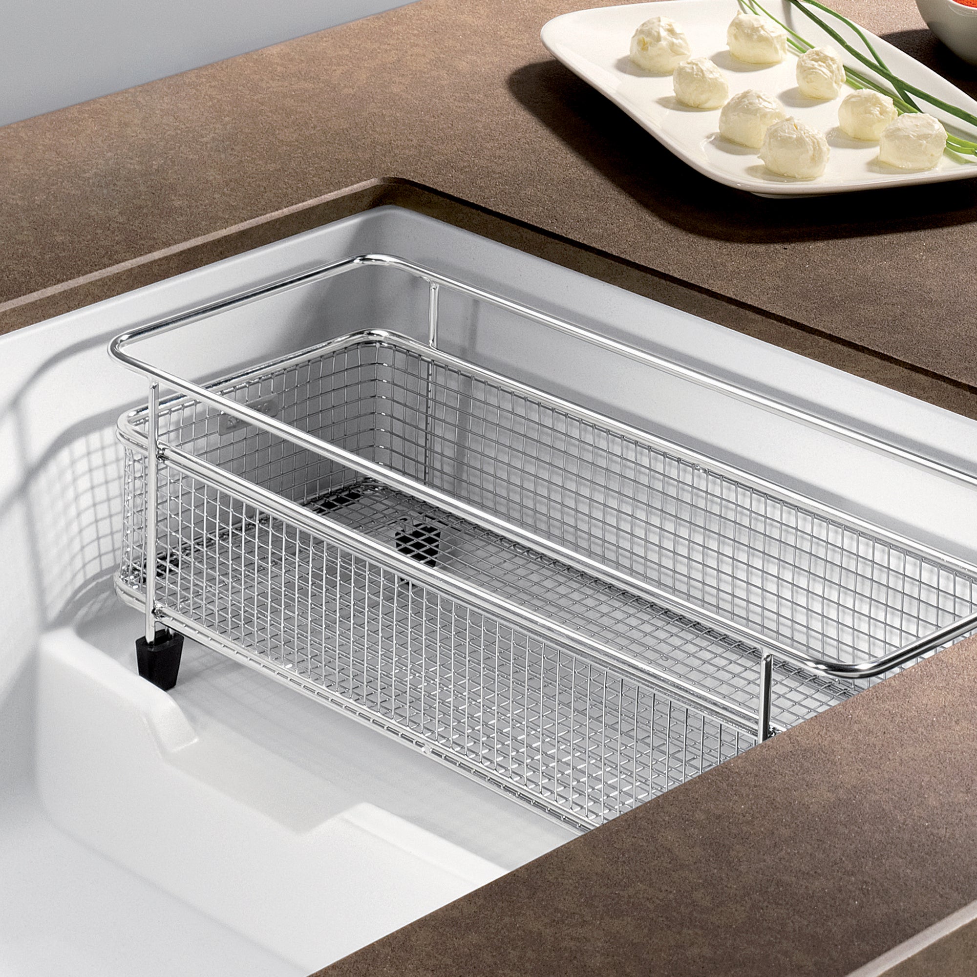 White undermount kitchen sink with metal strainer basket installed in countertop, adjacent to plate with appetizers.