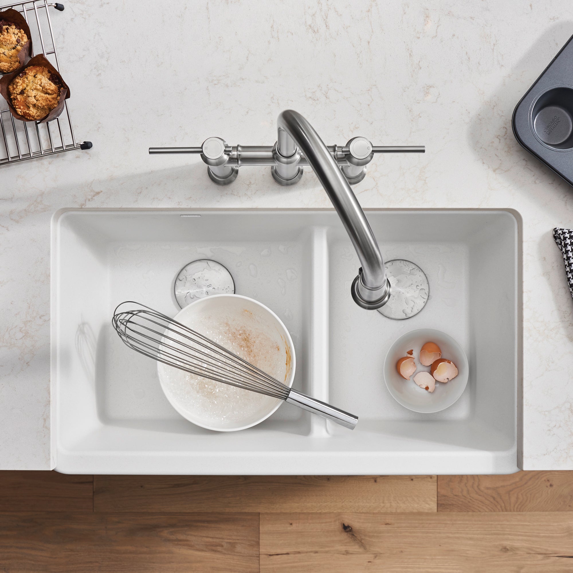 White double bowl apron sink with low divide, stainless steel faucet, and bowls containing mixing bowl and eggshells.