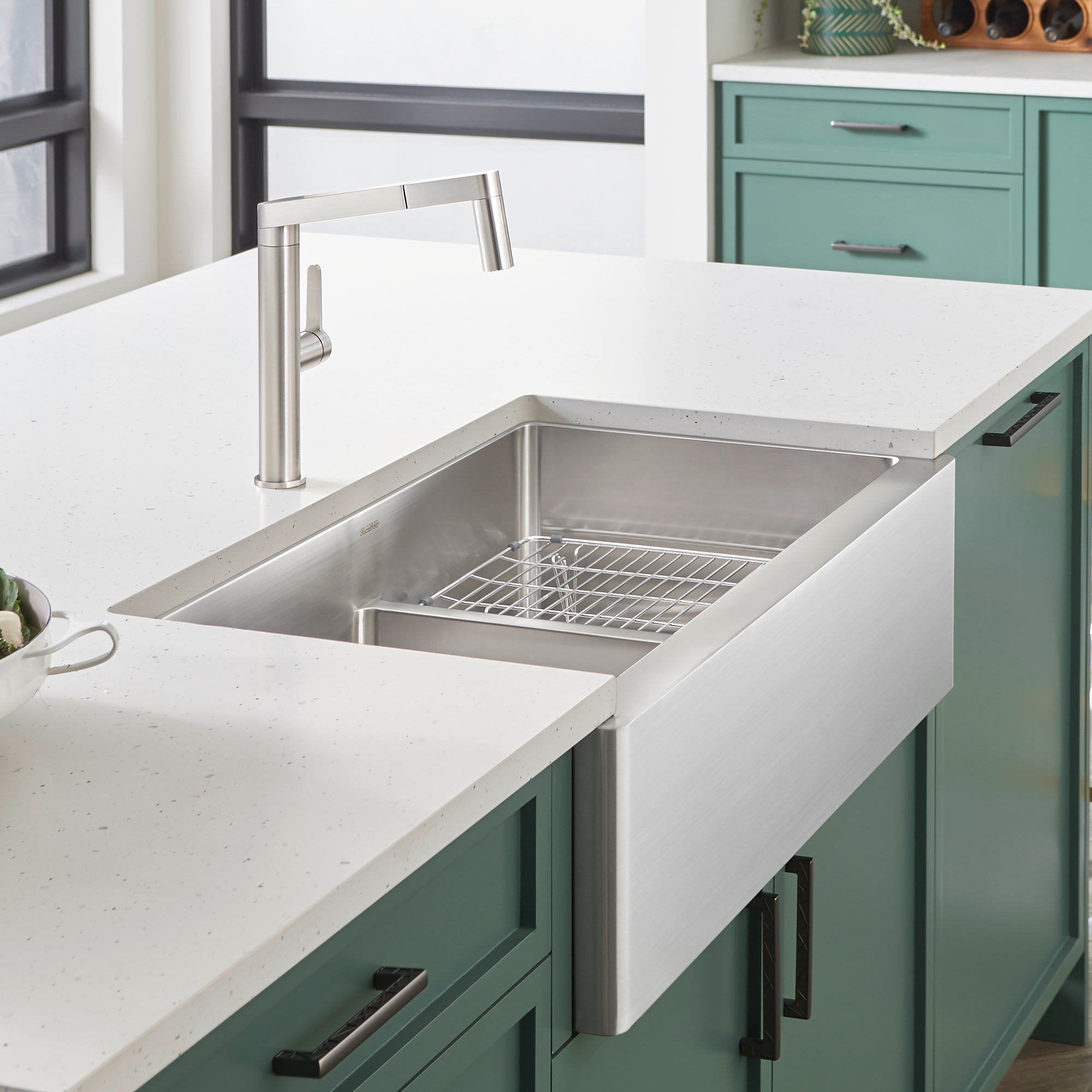 Stainless steel apron sink with low divider and metal grid insert installed in white countertop with faucet.