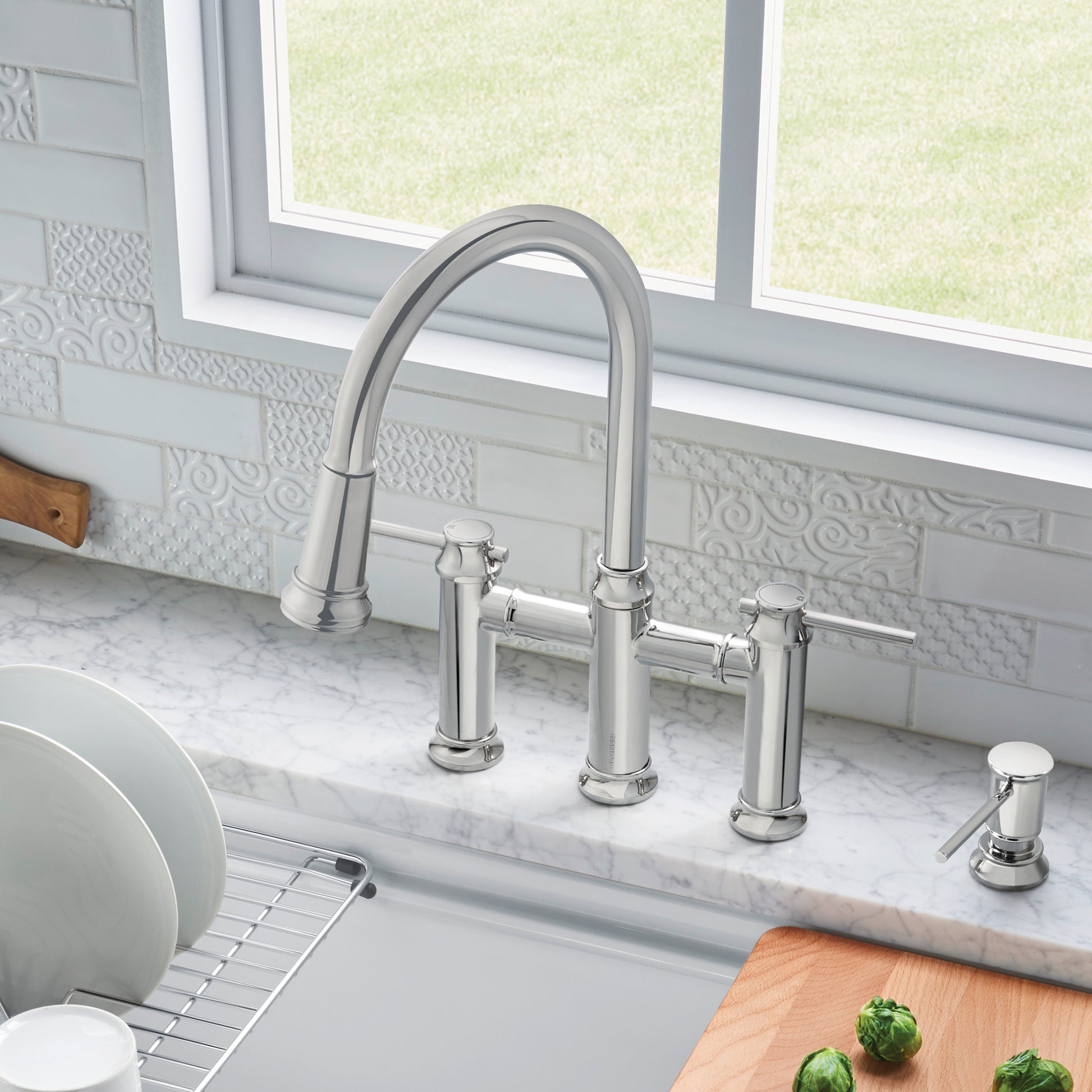 Chrome bridge pull-down faucet with curved spout and dual handles installed on marble countertop.