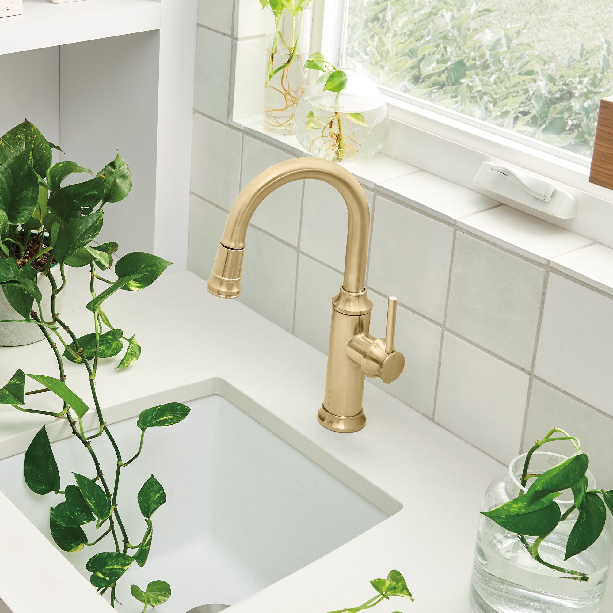 Satin gold pull-down bar faucet with curved spout mounted on white sink counter next to potted plants.