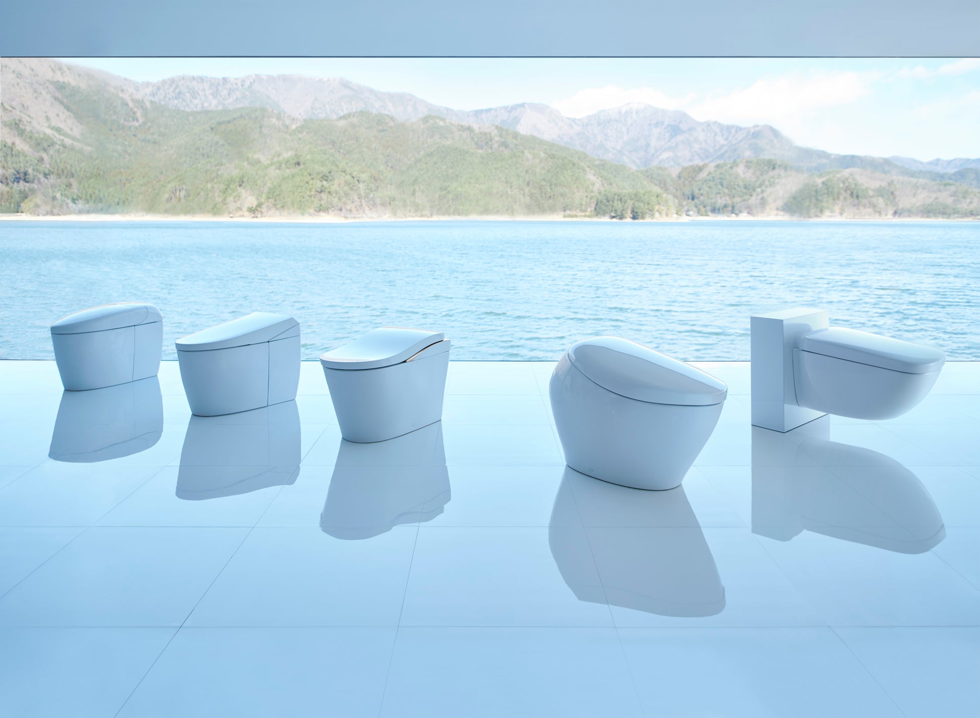 Five white wall-hung toilets with integrated bidet seats displayed in a row against a lakeside mountain backdrop.