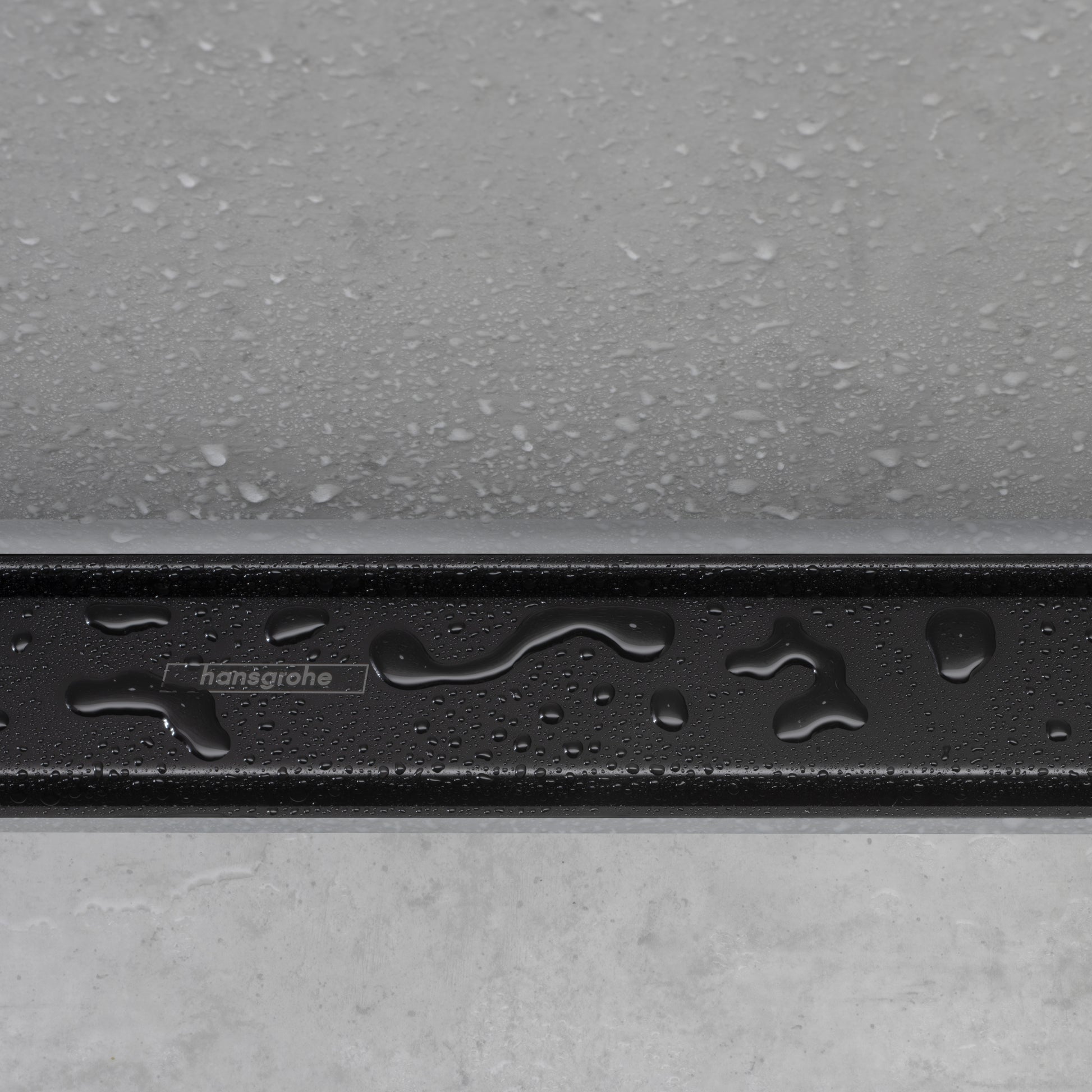 Matte black drain trim with hansgrohe branding, water droplets on surface, installed between gray tile and concrete.