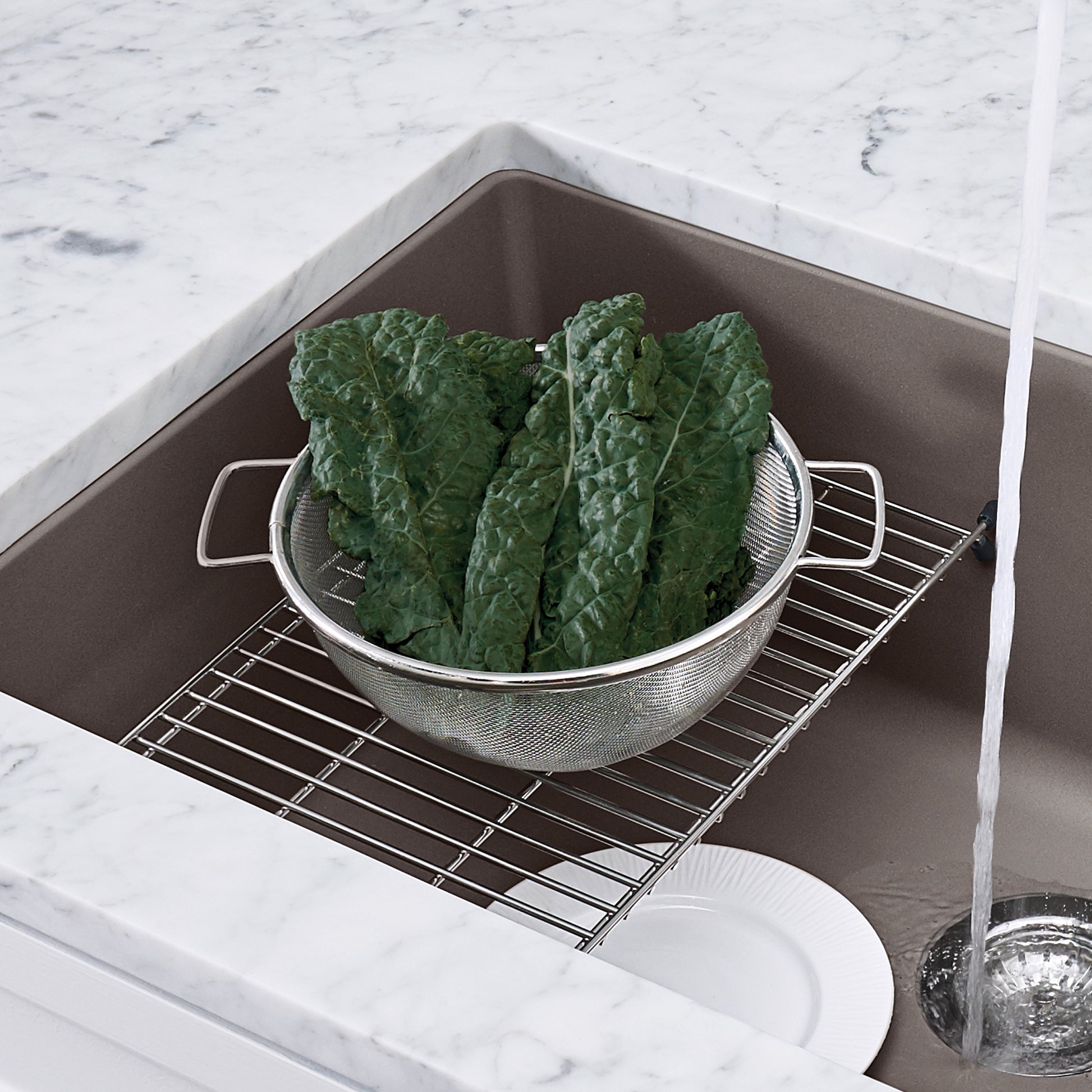 Stainless steel sink grid with handles resting in dark gray basin, supporting colander of fresh greens.