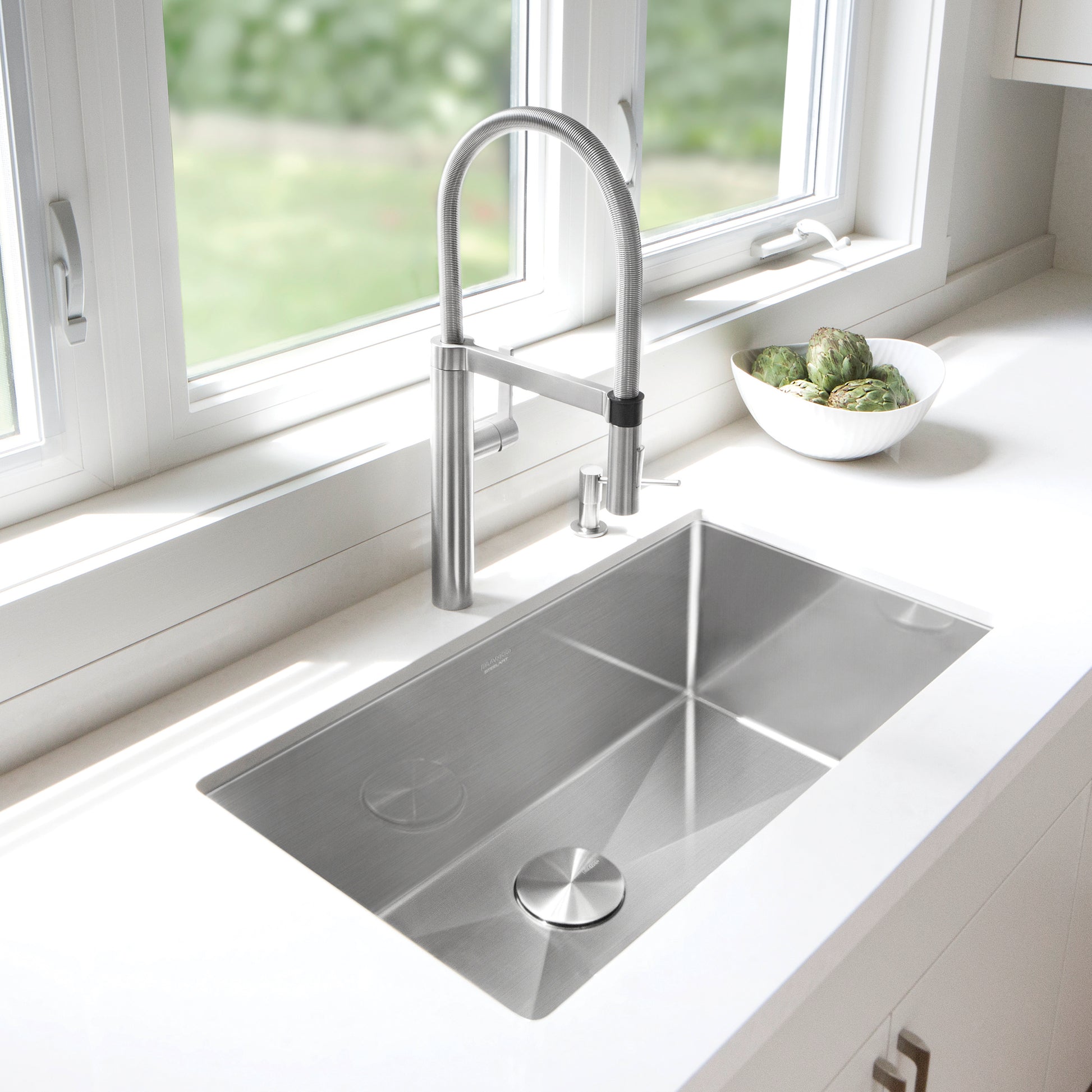 Stainless steel undermount kitchen sink with single bowl and curved pull-down faucet installed in white countertop.