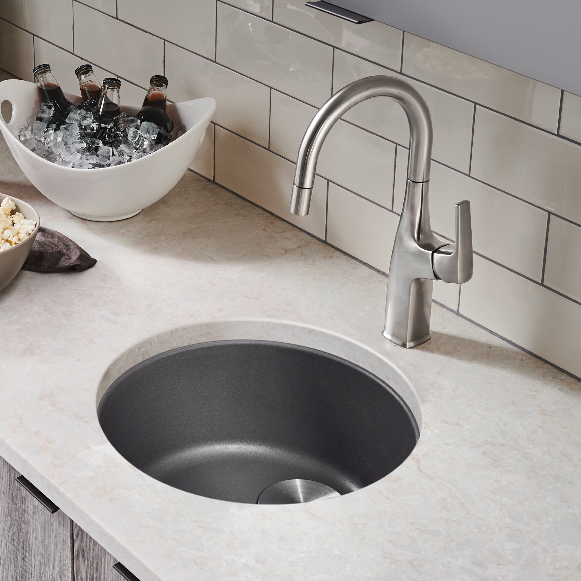Cinder-colored round bar sink with stainless steel faucet mounted on countertop with white subway tile backsplash.