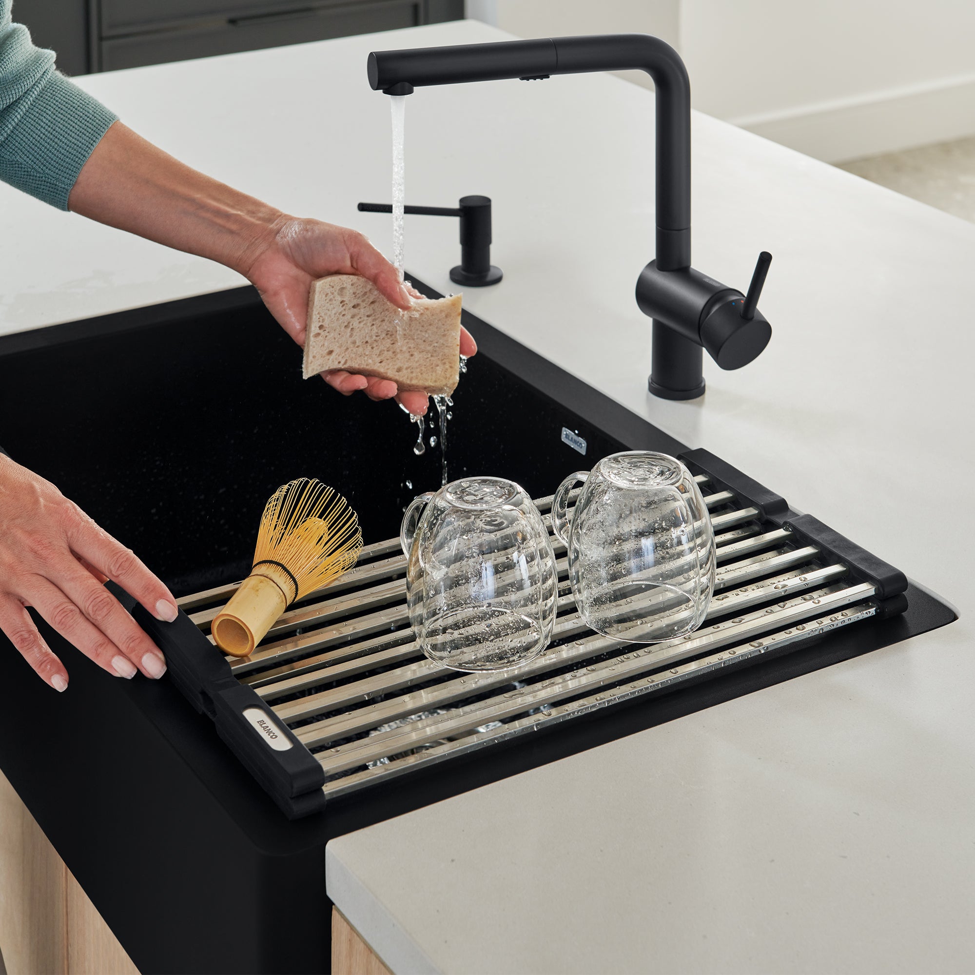 Black foldable drying mat with metal slats holding wet glasses and a brush next to a kitchen sink faucet.