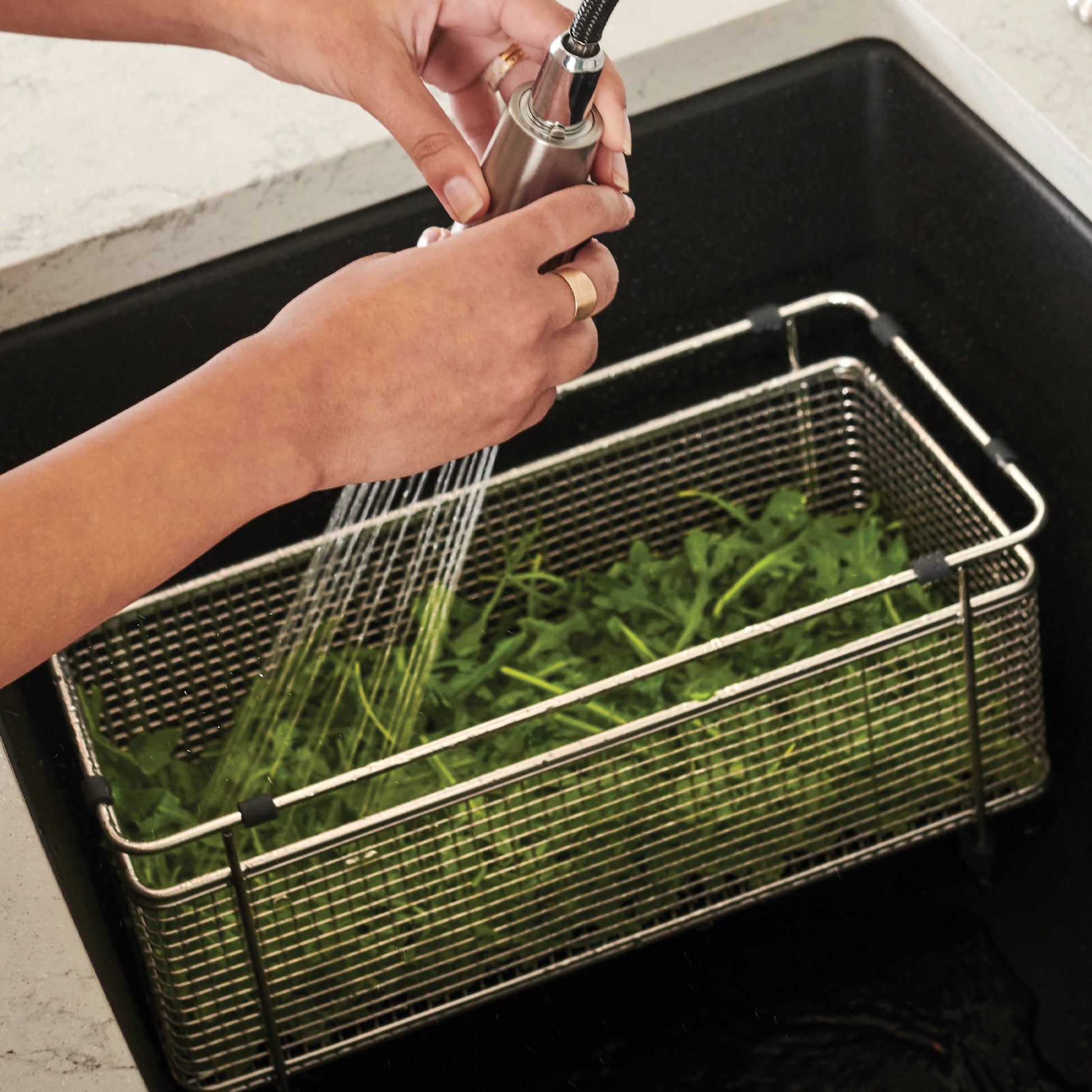 Stainless steel mesh colander with handles being rinsed under faucet while containing fresh herbs.