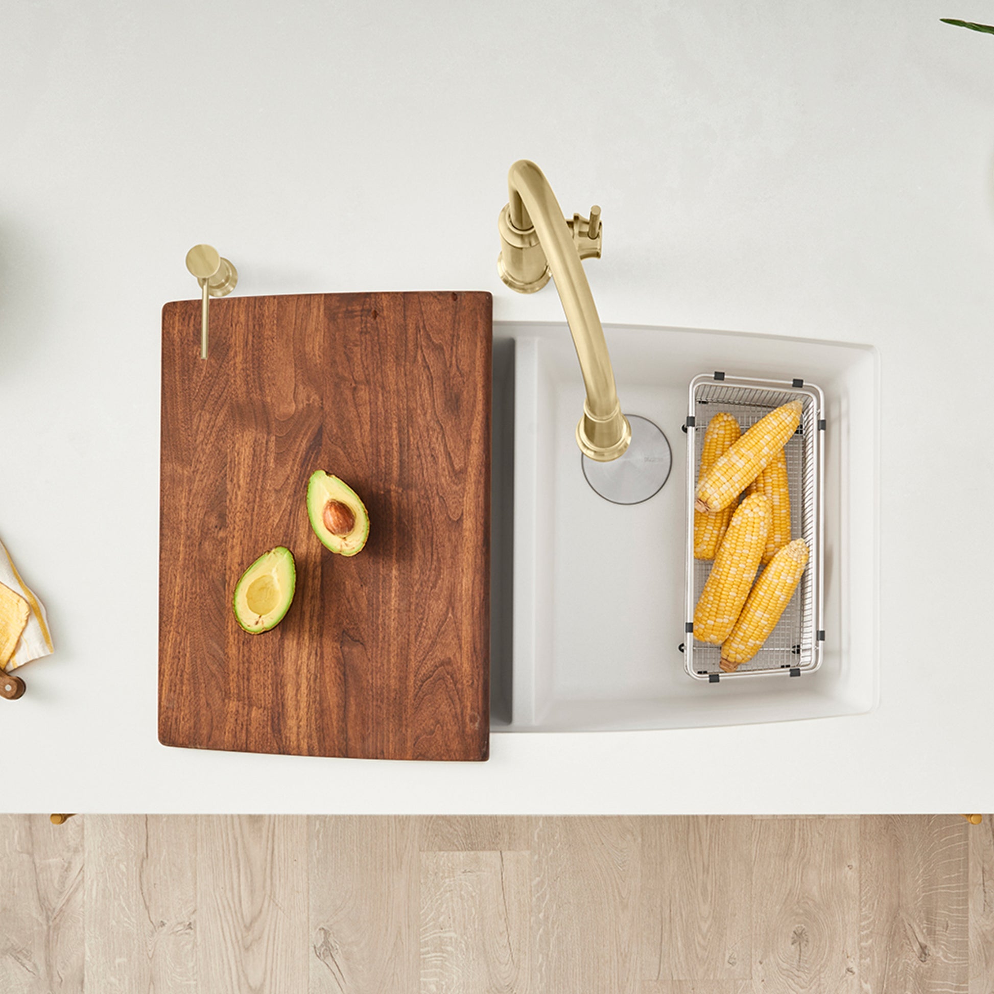 Brown wood cutting board with avocado halves, positioned over white double bowl sink with brass faucet and colander insert.