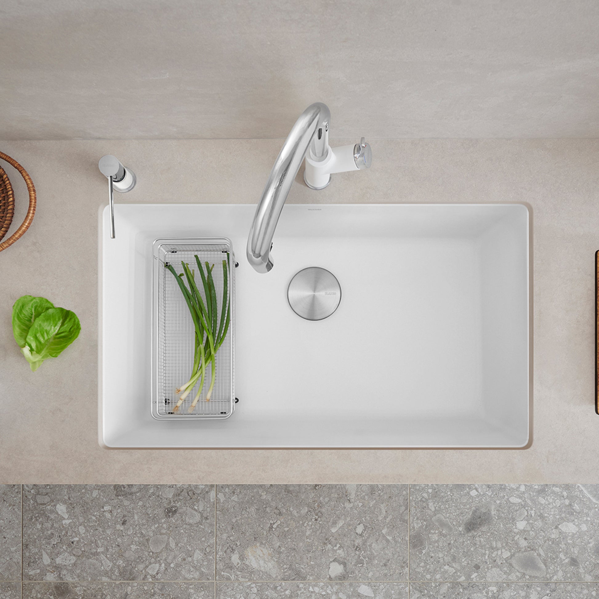 Stainless steel mesh basket with fresh herbs inside, positioned in a white kitchen sink.