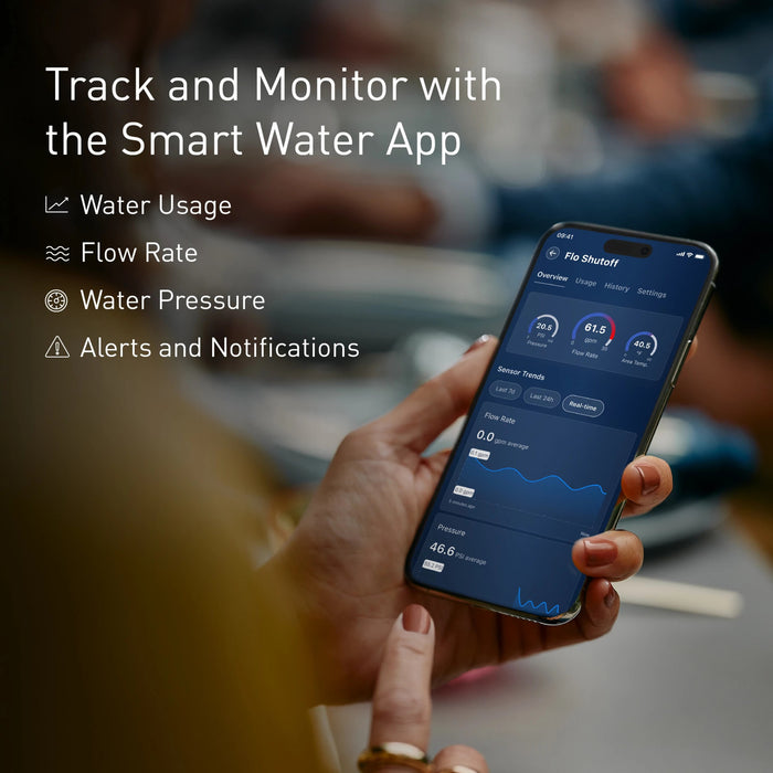 Person holding smartphone displaying Moen Flo water monitoring app with usage, flow rate, and pressure data on screen.