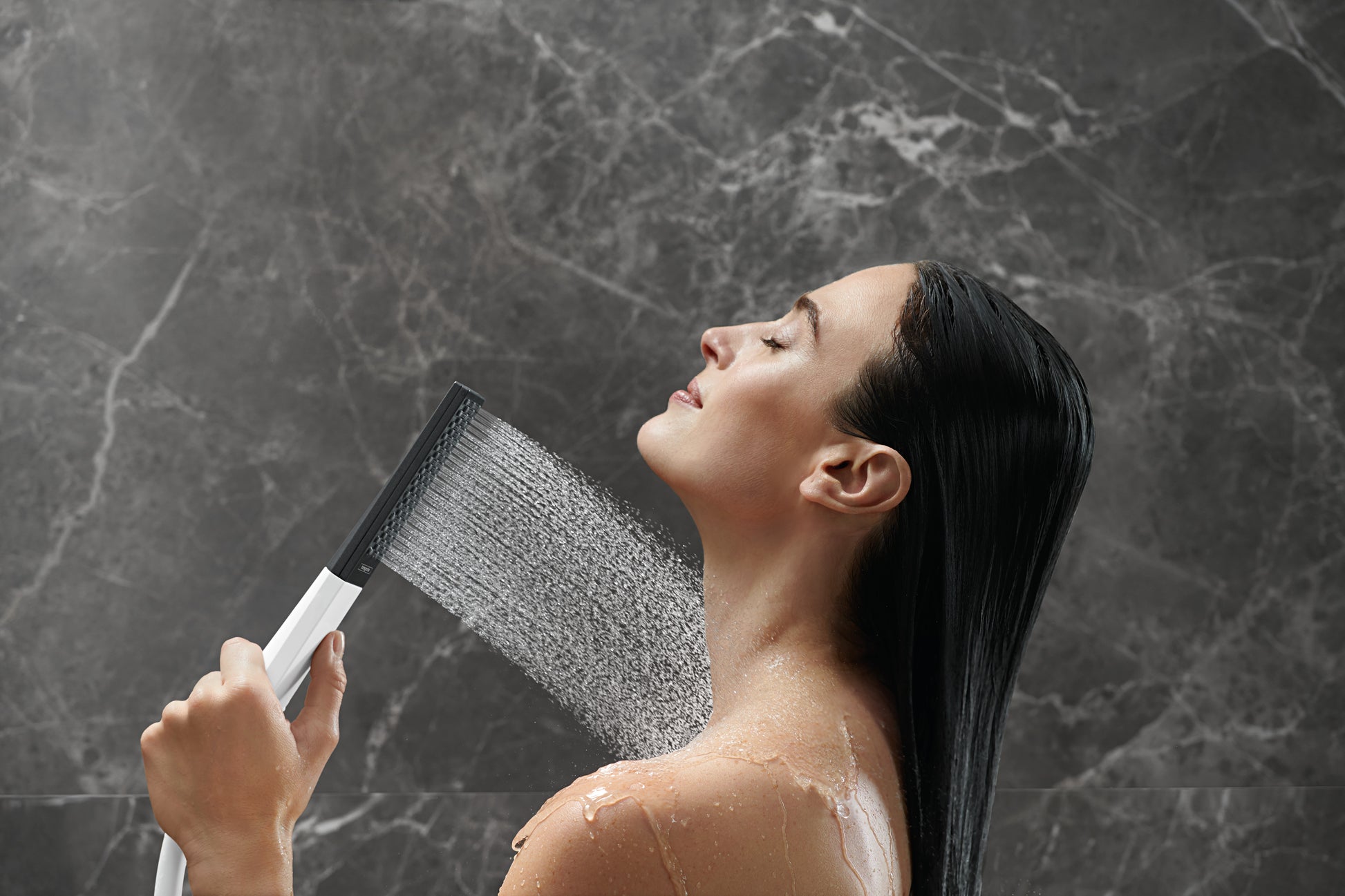Chrome handheld shower head with rectangular face spraying water, held by person under running water against marble tile back